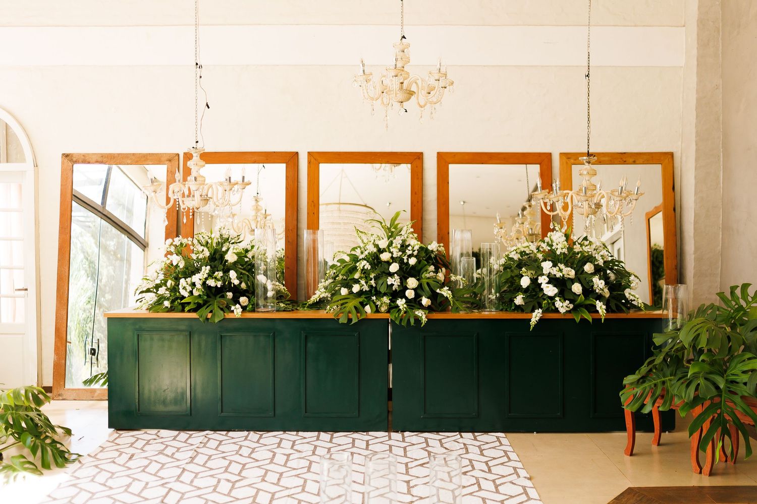 Elegant reception desk adorned with lush white floral arrangements, gold-framed mirrors, and crystal chandeliers in a bright venue.
