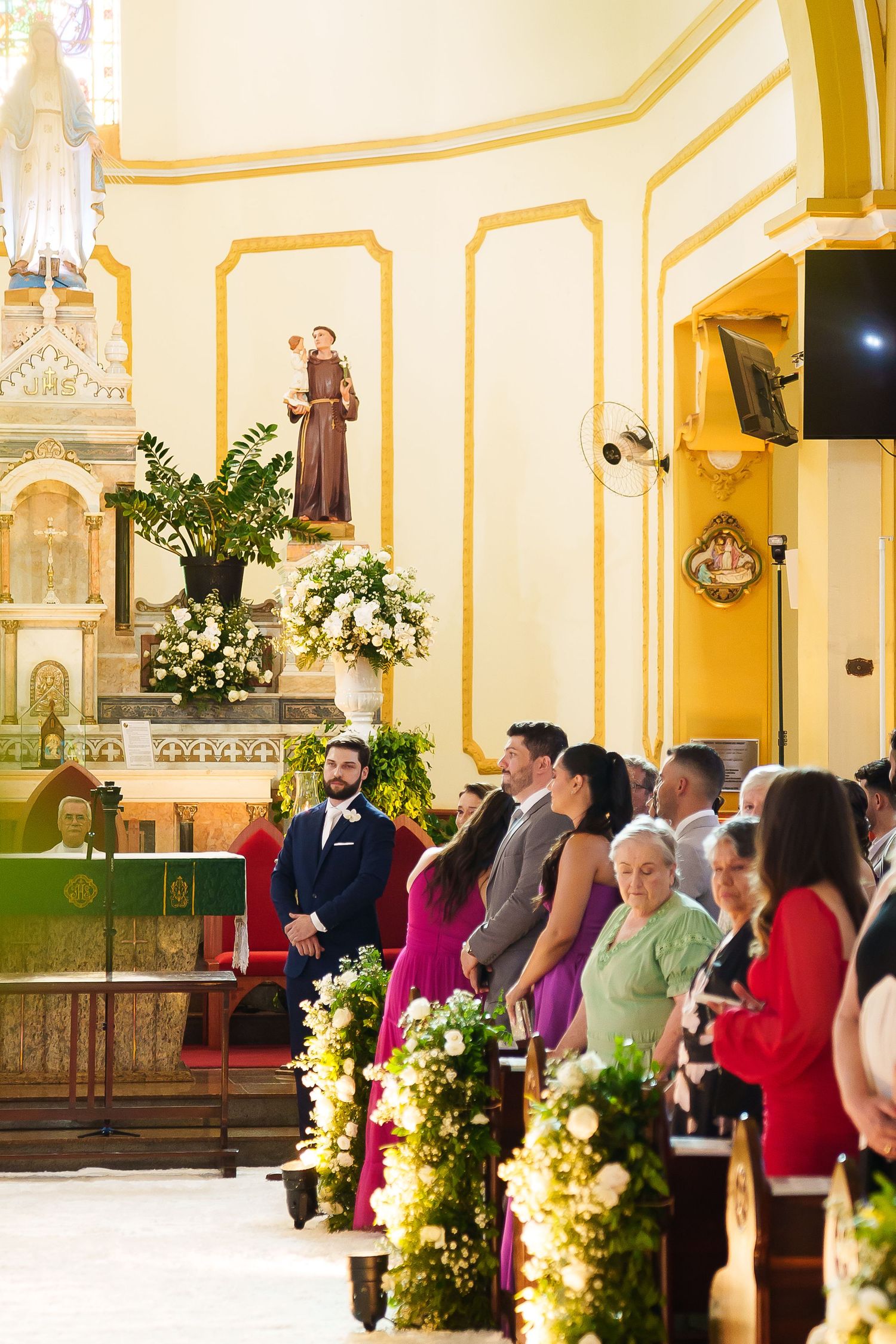 Groom stands at church altar during wedding ceremony with floral arrangements and guests seated in pews.