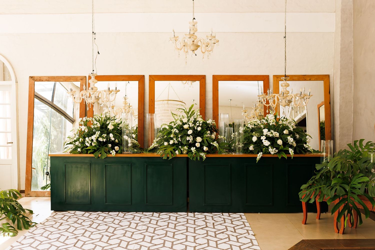 Elegant wedding reception bar with green paneling, gold mirrors, crystal chandeliers, and lush white floral arrangements.