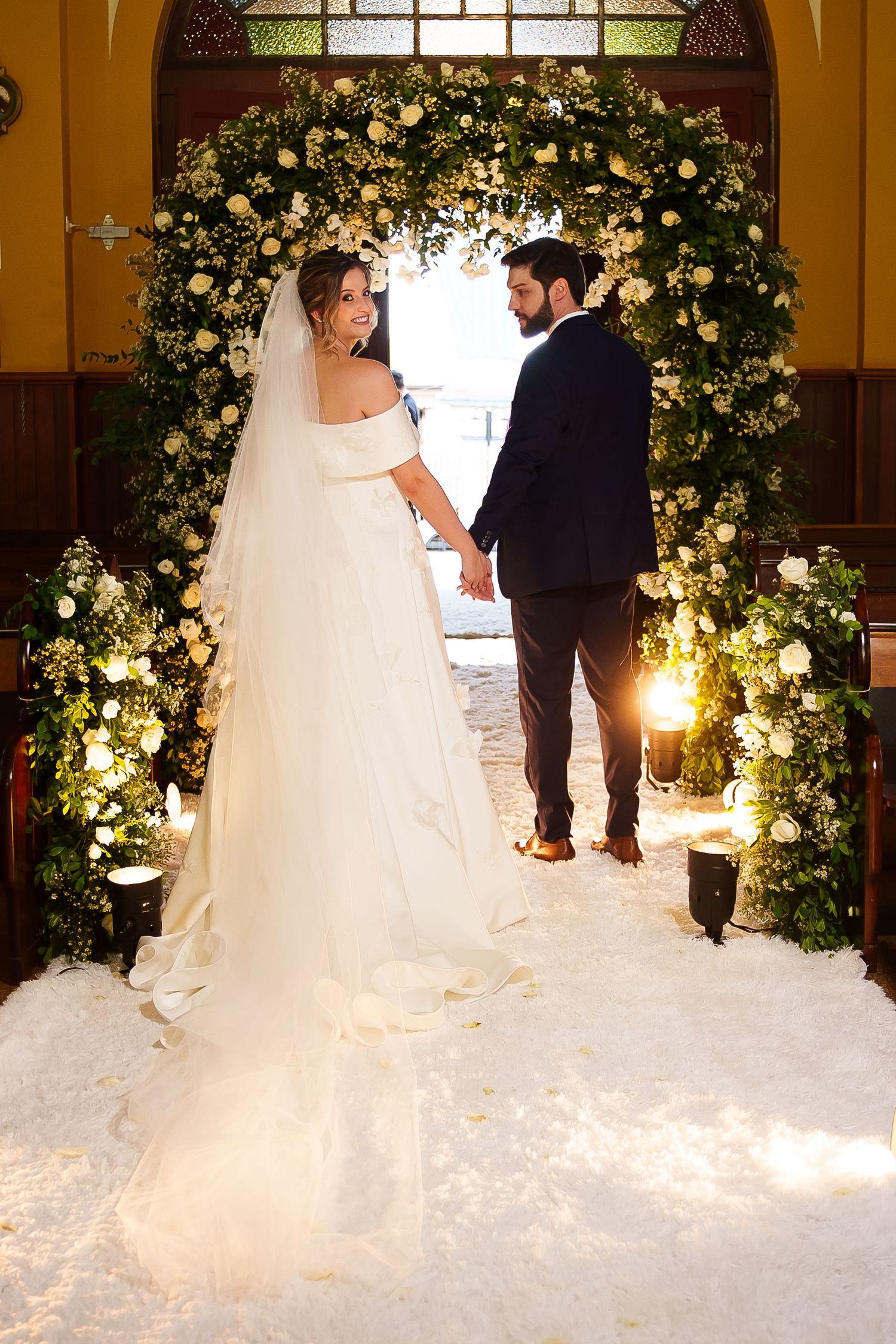 Bride and groom holding hands at floral arch altar in church, white gown with veil, groom in dark suit.