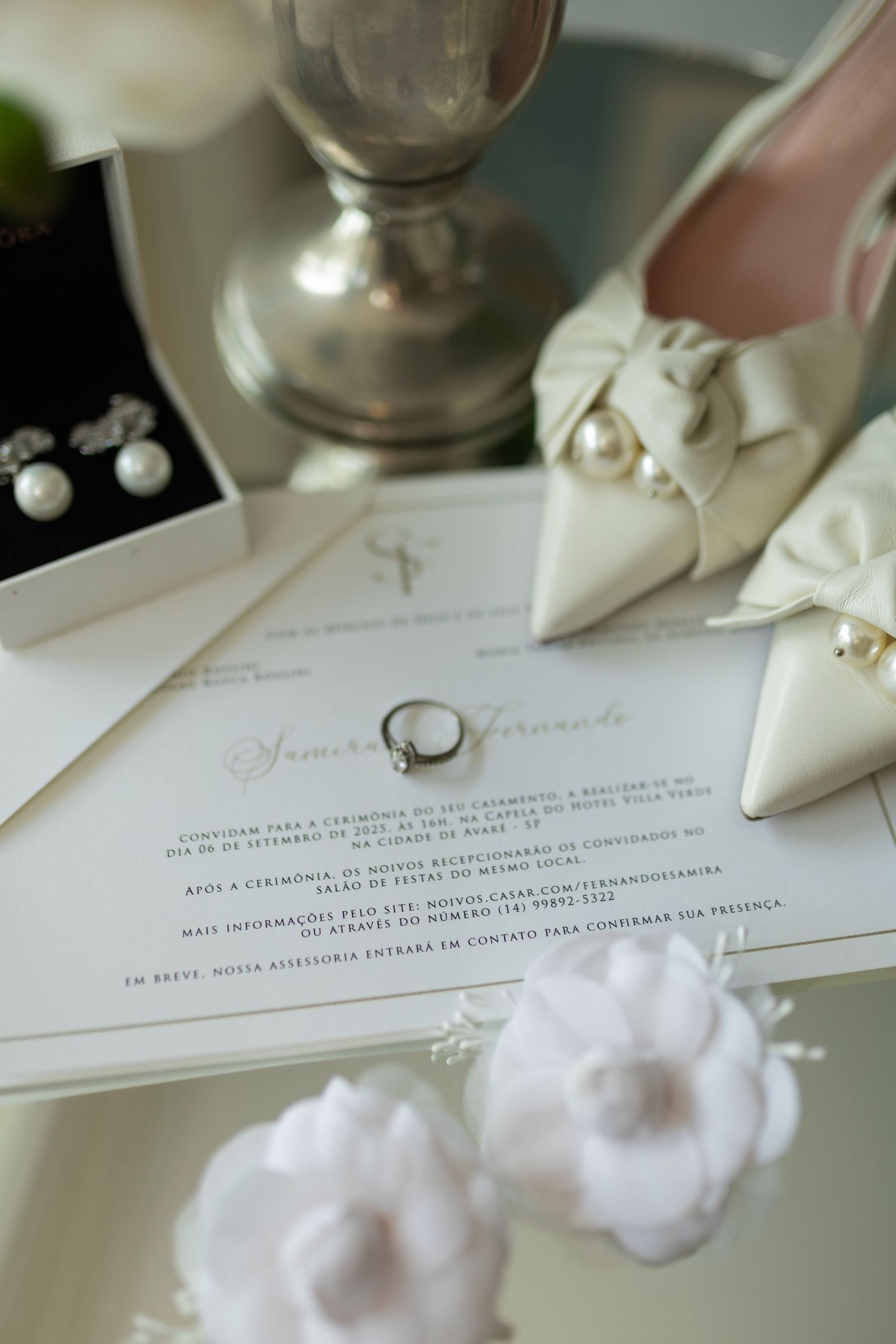 Elegant wedding flat lay with diamond ring on invitation, pearl earrings, white bow heels, and floral accessories.