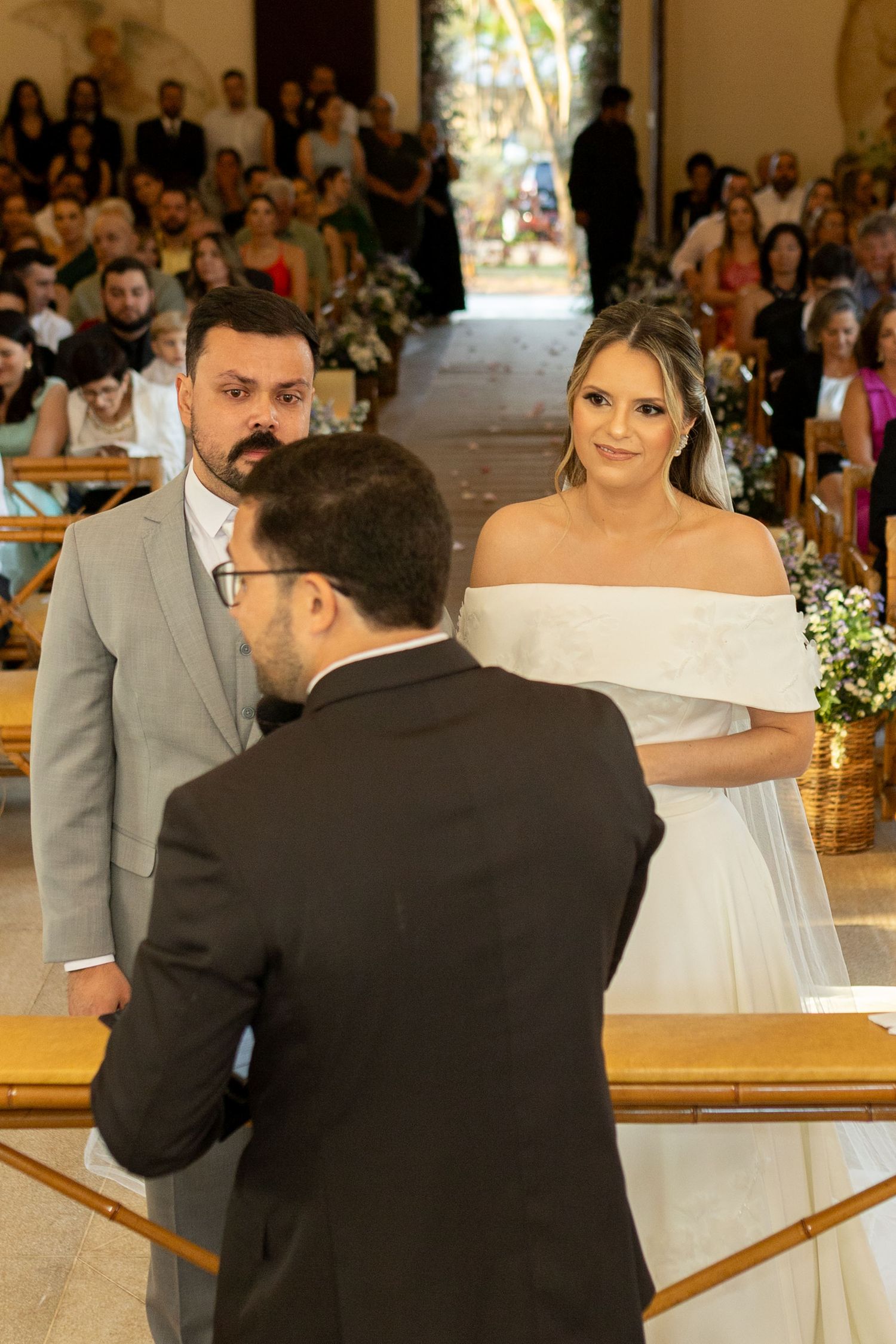 Bride in off-shoulder white gown and groom exchange vows at church altar with wedding guests seated in background.