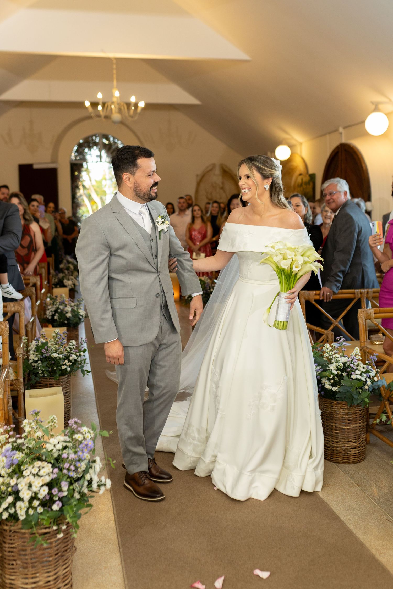 Bride in white off-shoulder gown holding calla lilies walks the aisle with groom in gray suit in a flower-decorated church.