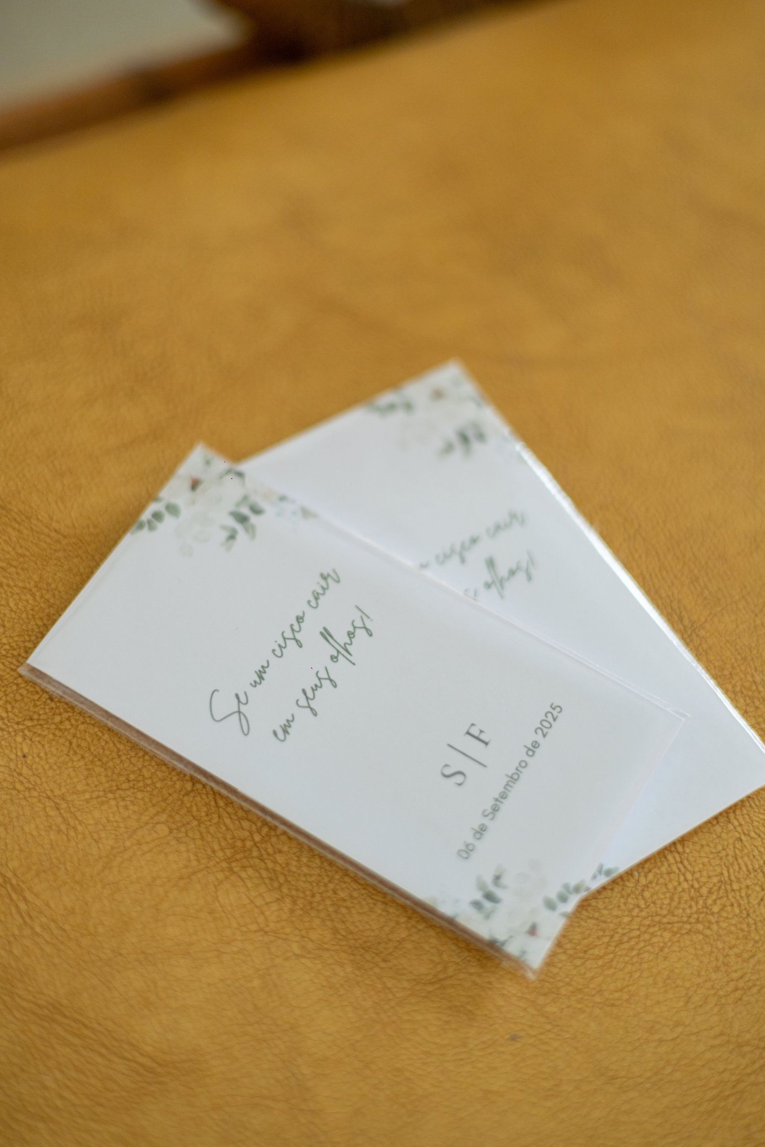 Two elegant white wedding invitation cards with green botanical details resting on a golden leather surface.