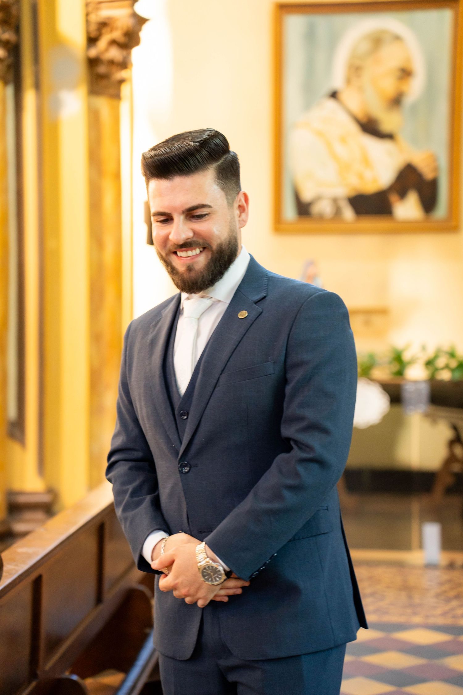 Smiling bearded man in navy blue three-piece suit standing in a church with a religious painting behind him.