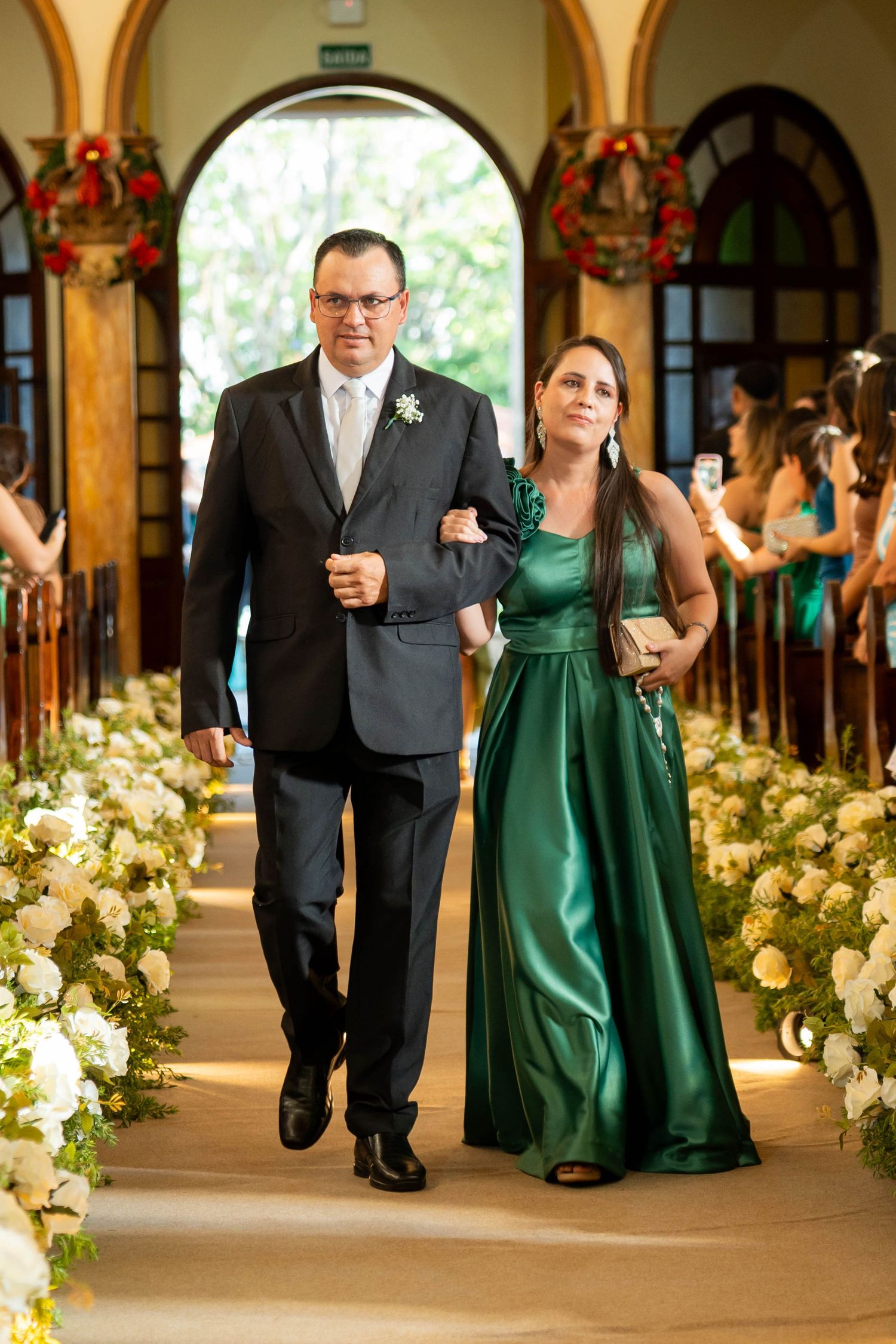 Couple walking down a flower-lined church aisle, man in gray suit, woman in elegant green satin gown.
