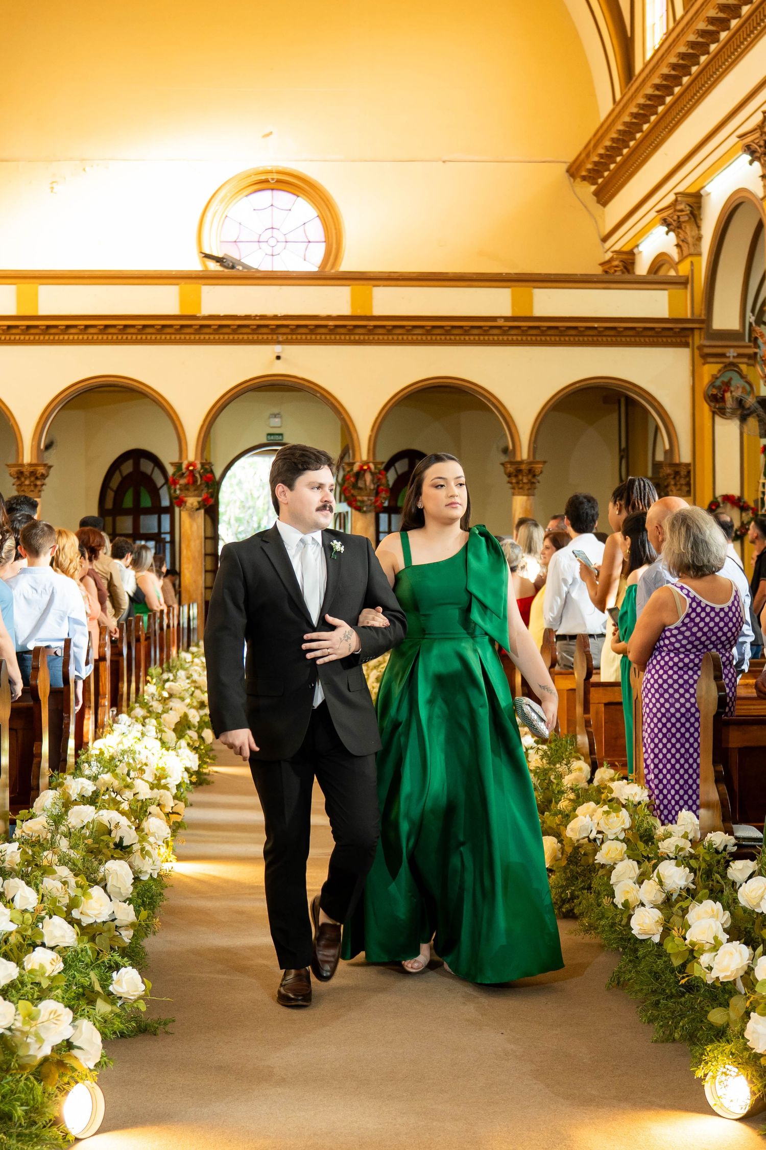 A man in a suit escorts a woman in an emerald green gown down a flower-lined church aisle at a wedding.