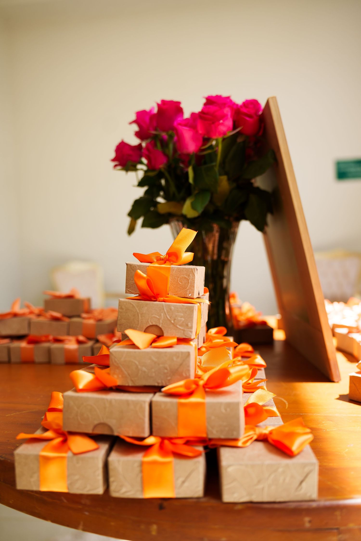 Stack of gold gift boxes with orange ribbons on a wooden table, pink roses bouquet in background.