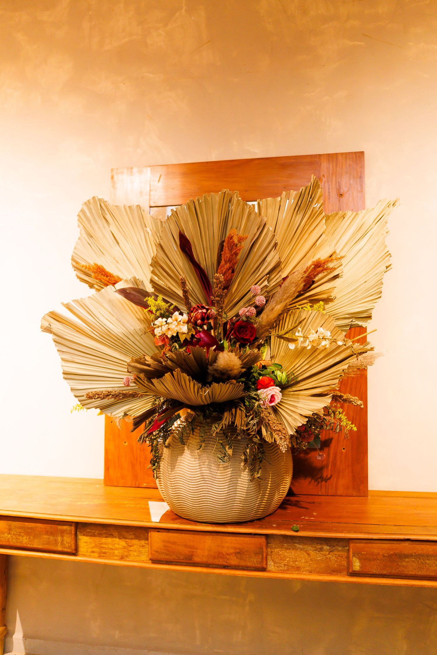 Dried palm leaf floral arrangement in a ceramic vase on a wooden console table with a warm neutral background.