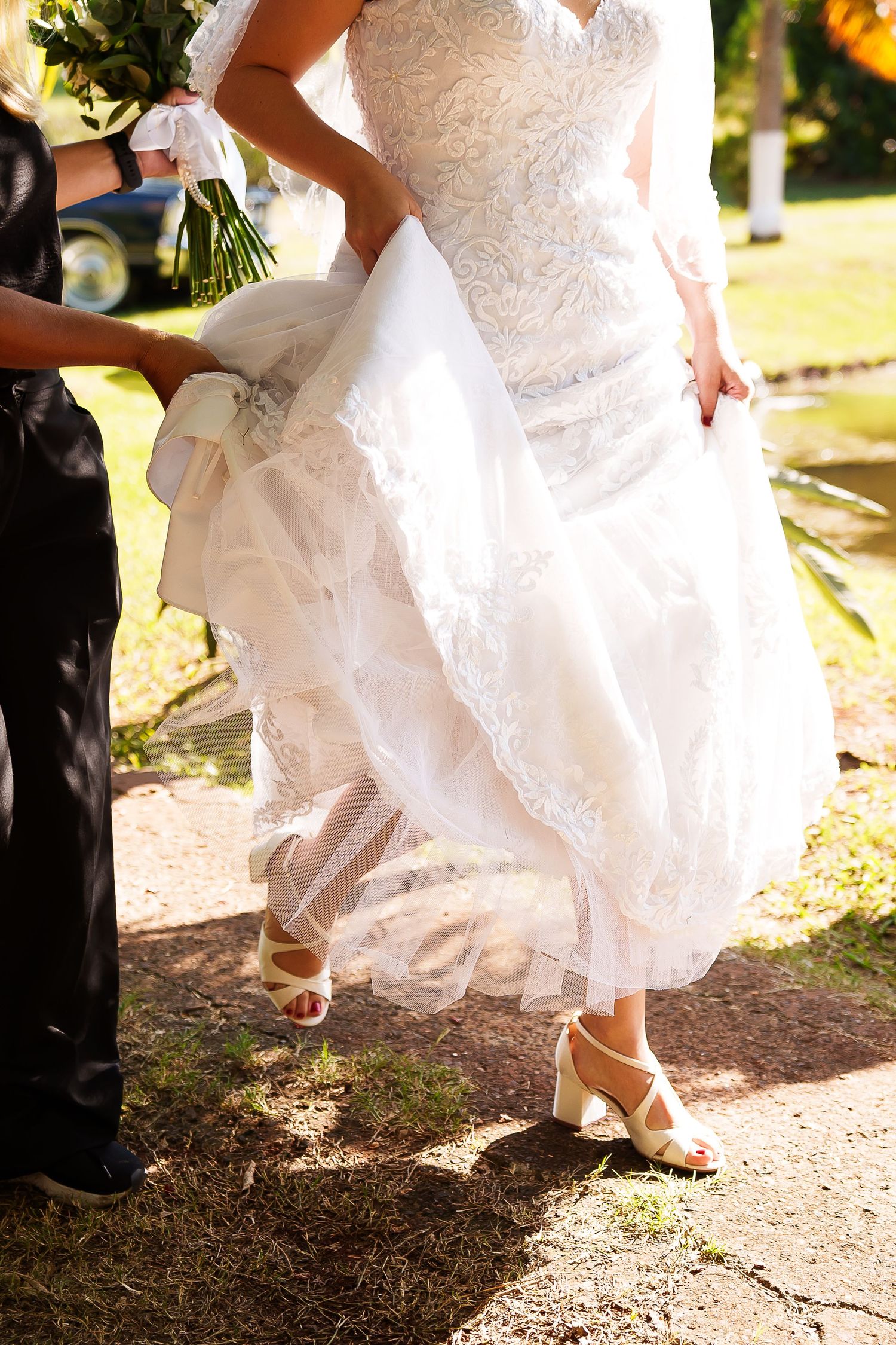 Bride in white lace wedding gown with tulle skirt walking outdoors, assisted by attendant, wearing ivory block heel shoes.