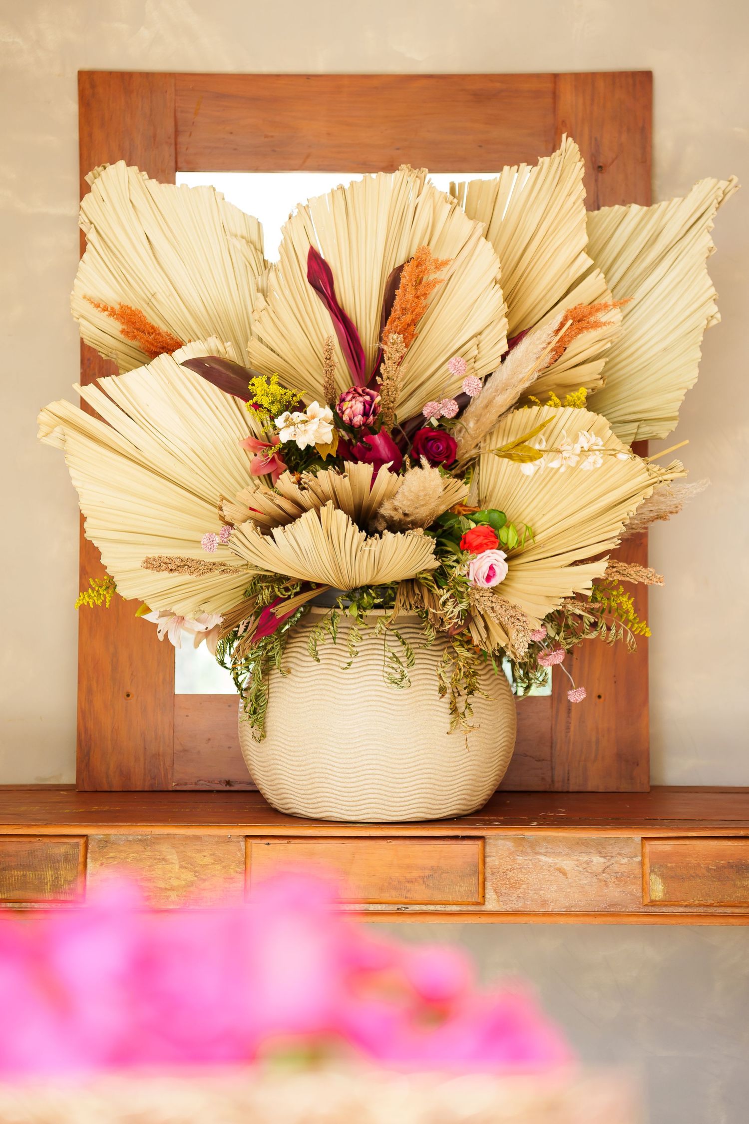 Boho dried palm leaf and wildflower arrangement in white ribbed vase displayed in front of wooden framed mirror.
