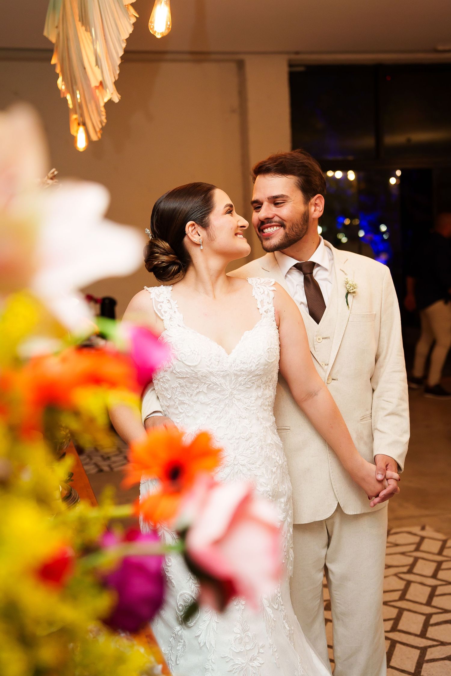 Bride in lace gown and groom in beige suit share a romantic moment at their wedding reception with colorful flowers.