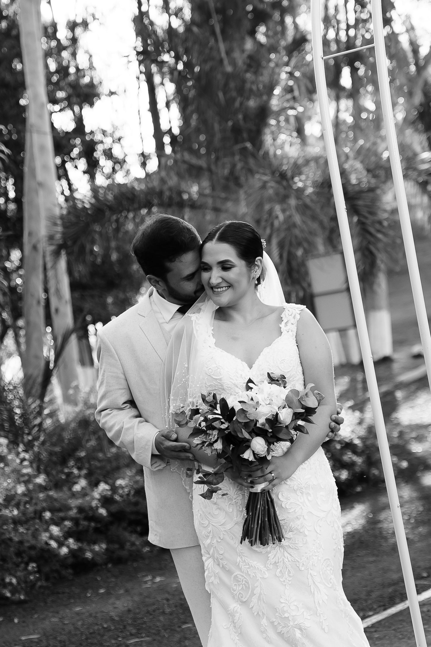 Black and white photo of a smiling bride holding bouquet as groom tenderly kisses her cheek outdoors.