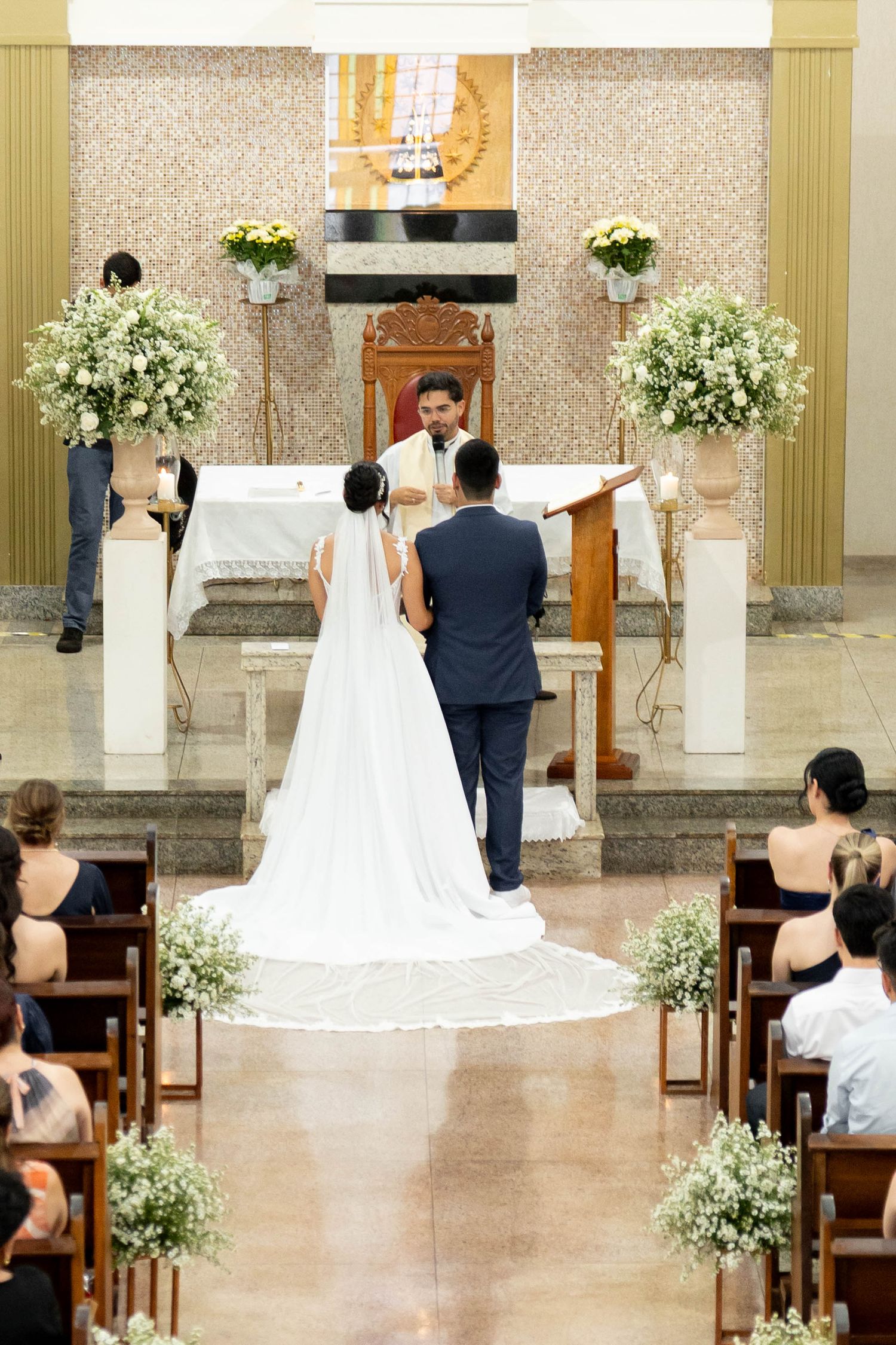Bride and groom standing at church altar during wedding ceremony with floral decorations and seated guests.