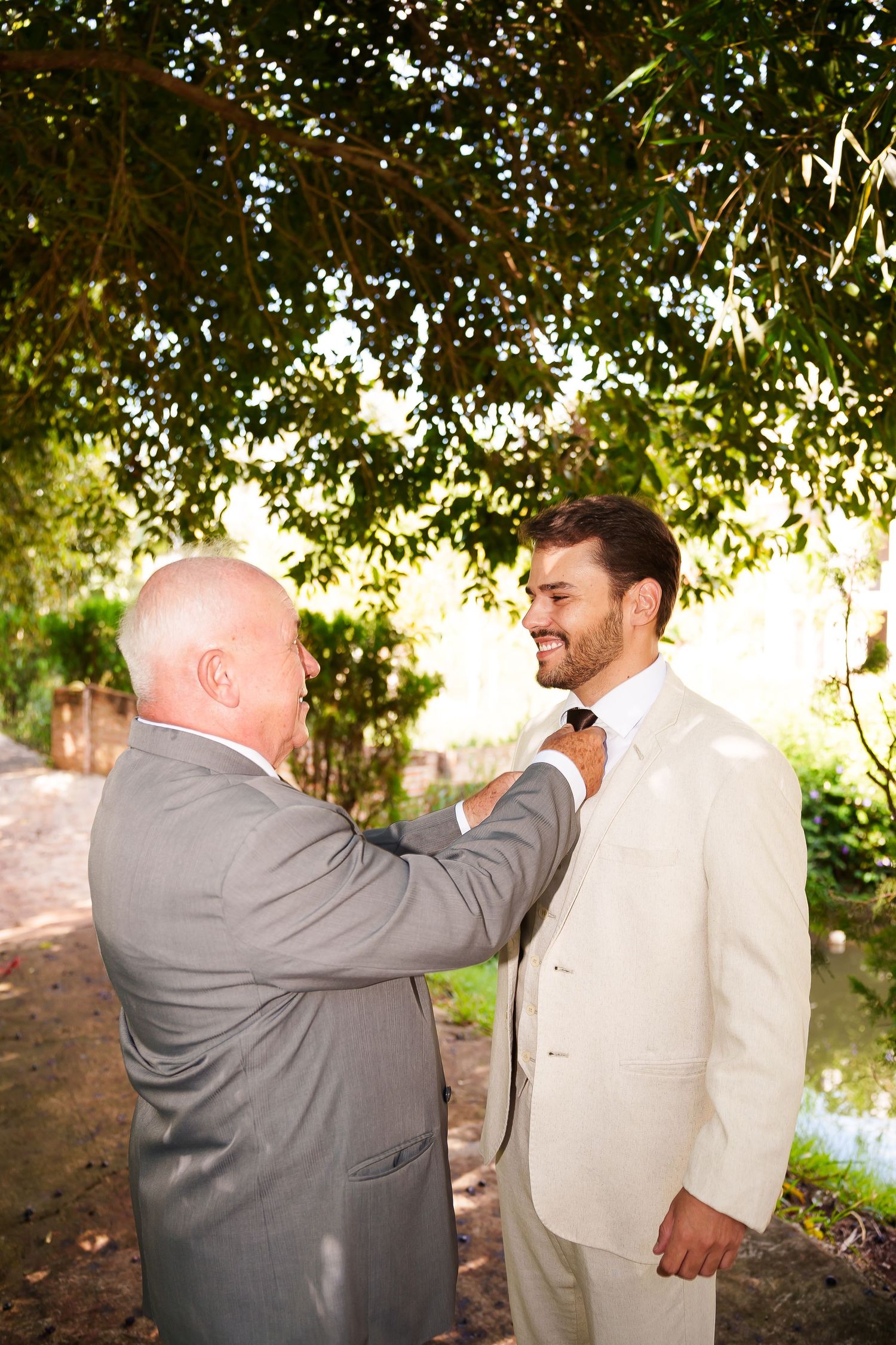 Two men in suits share a joyful moment outdoors, with one adjusting the other's tie before a wedding ceremony.