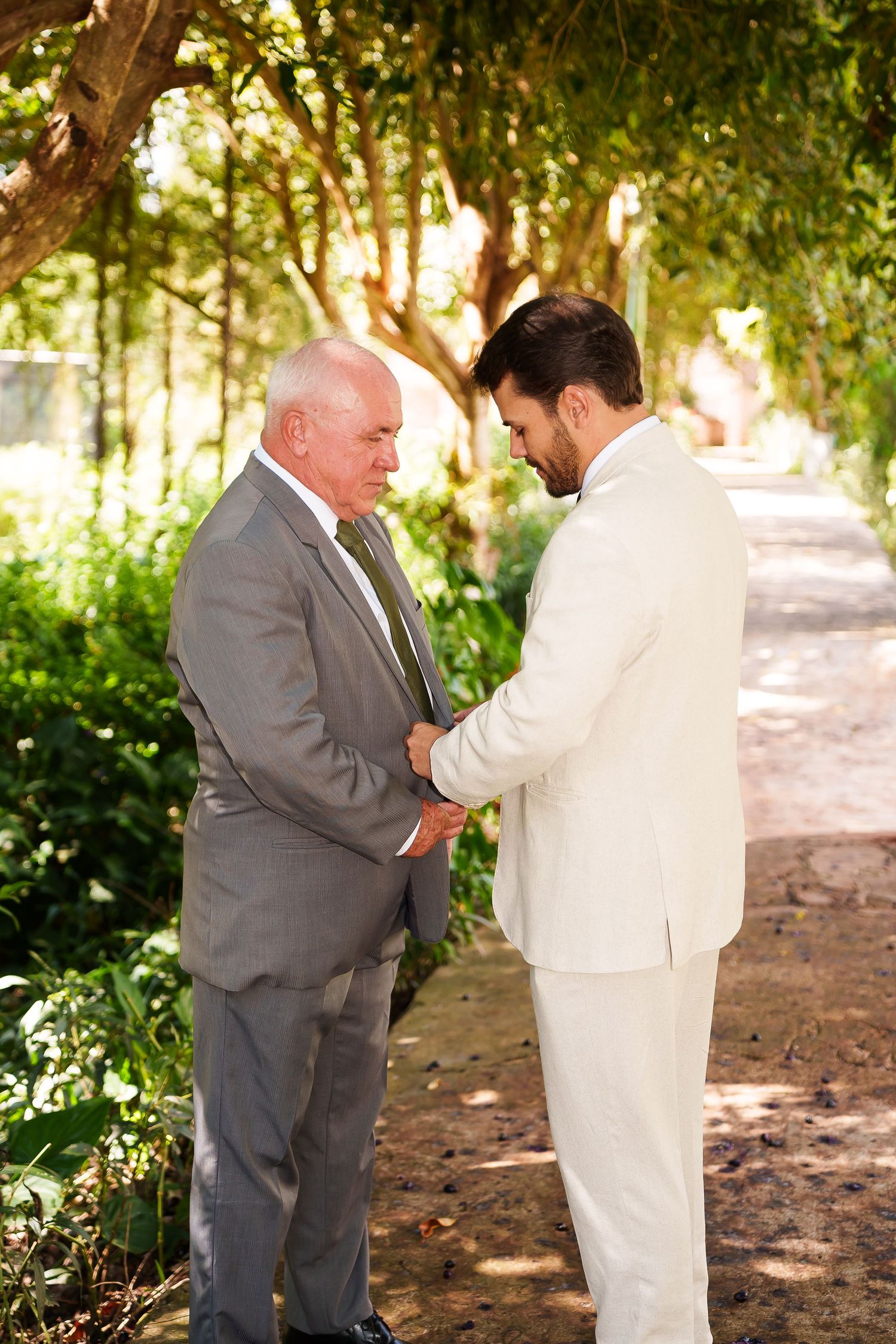 Two men in suits share a tender moment outdoors, with lush green trees in the background at a wedding.