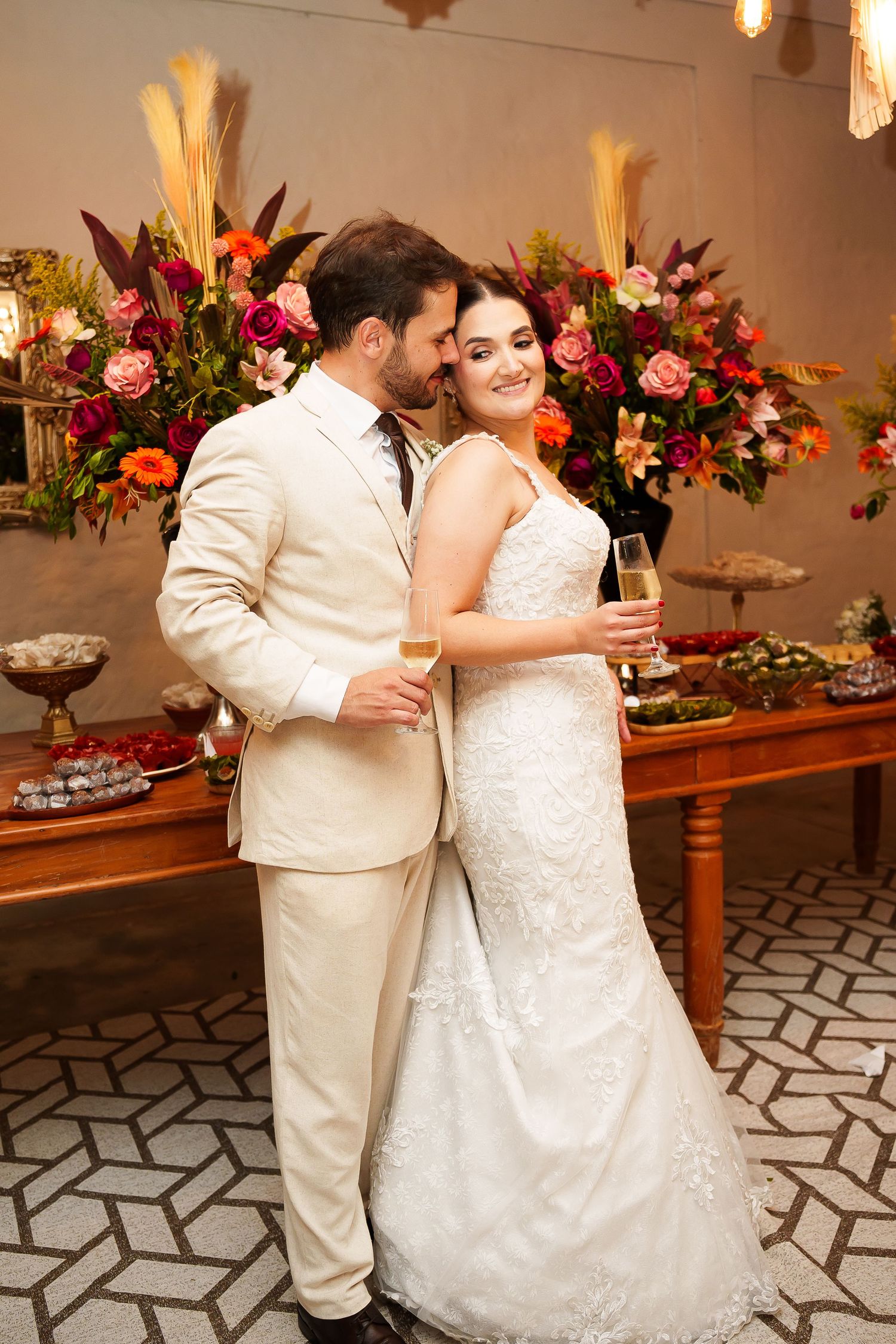 Bride and groom holding champagne glasses pose together in front of colorful floral arrangements at their wedding reception.