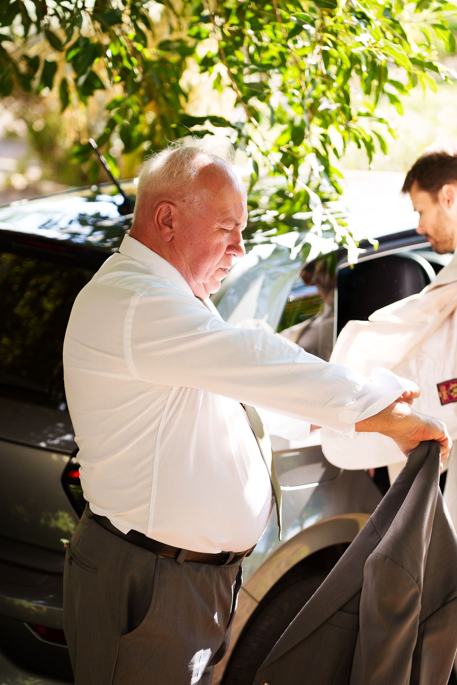 Elderly man in white shirt holding a suit jacket near a car outdoors on a sunny day.