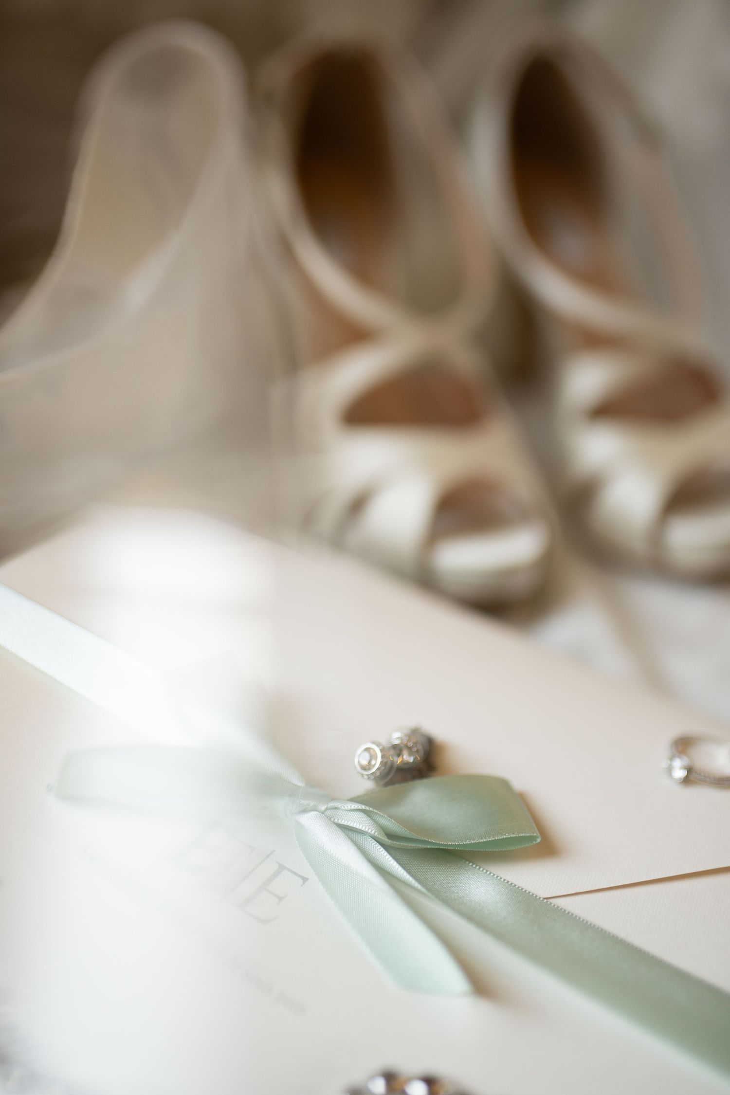 Elegant bridal flat lay with sage green ribbon, diamond ring, pearl earrings, and ivory heels on white background.