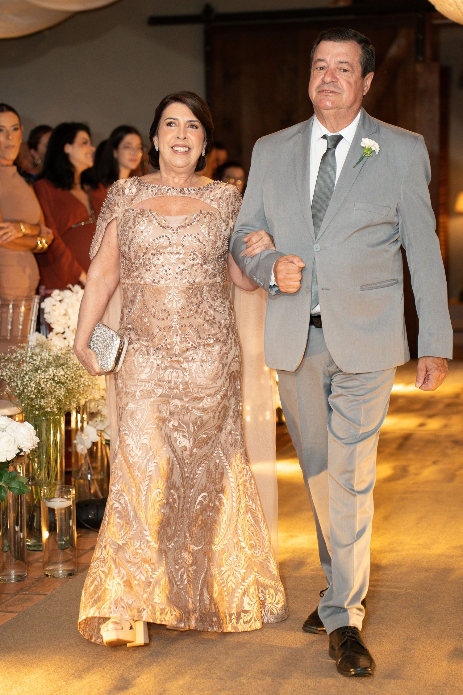 Elegant couple walking down wedding aisle, woman in gold gown, man in gray suit with boutonniere.