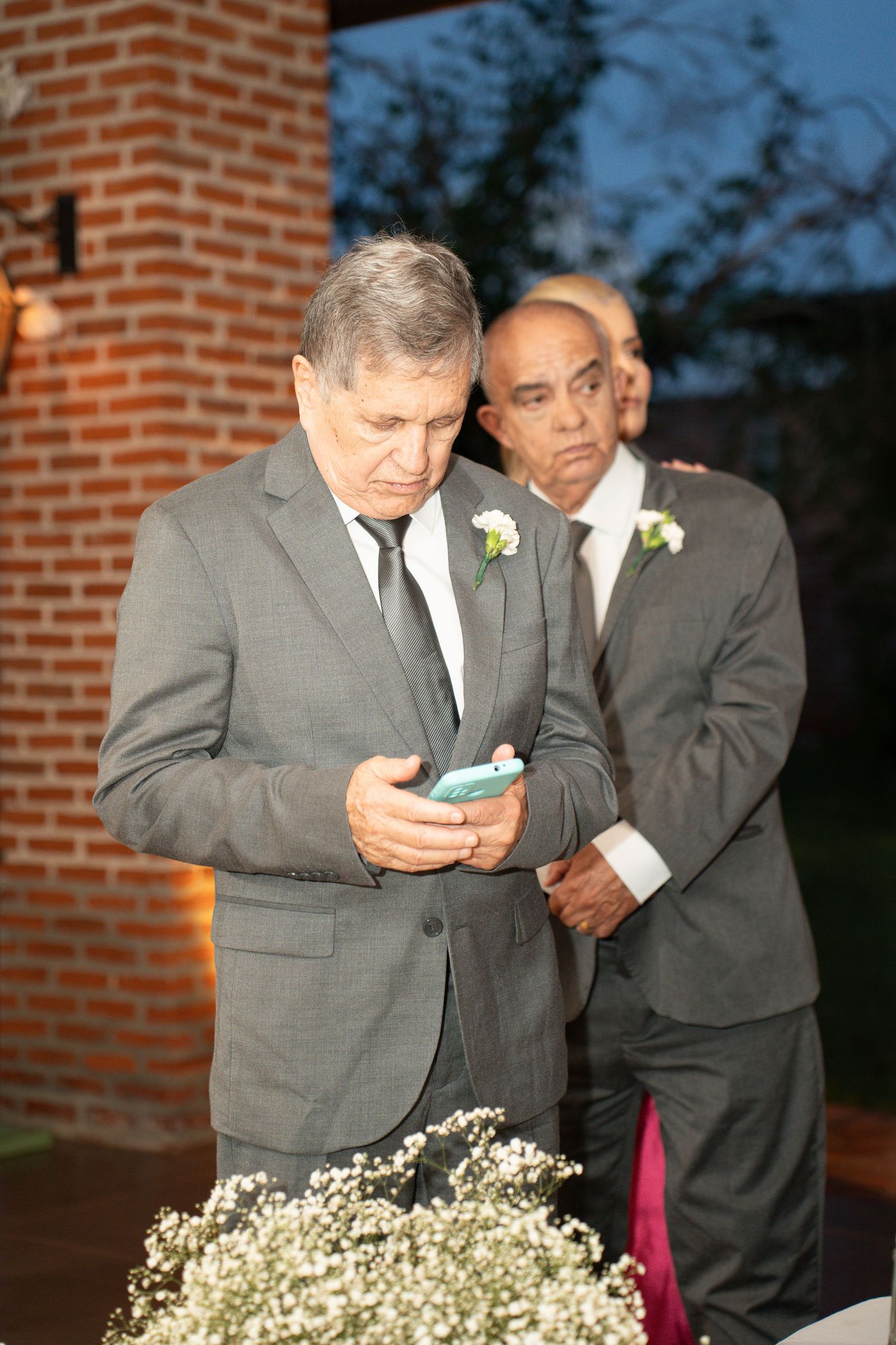 Two men in gray suits with boutonnieres at a formal event, one looking at a phone near white flowers.