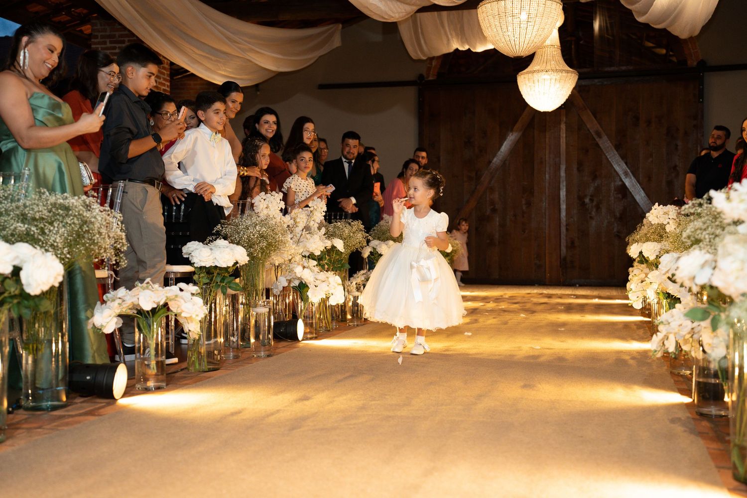 A flower girl in a white dress walks down a floral-lined wedding aisle as guests watch and celebrate.