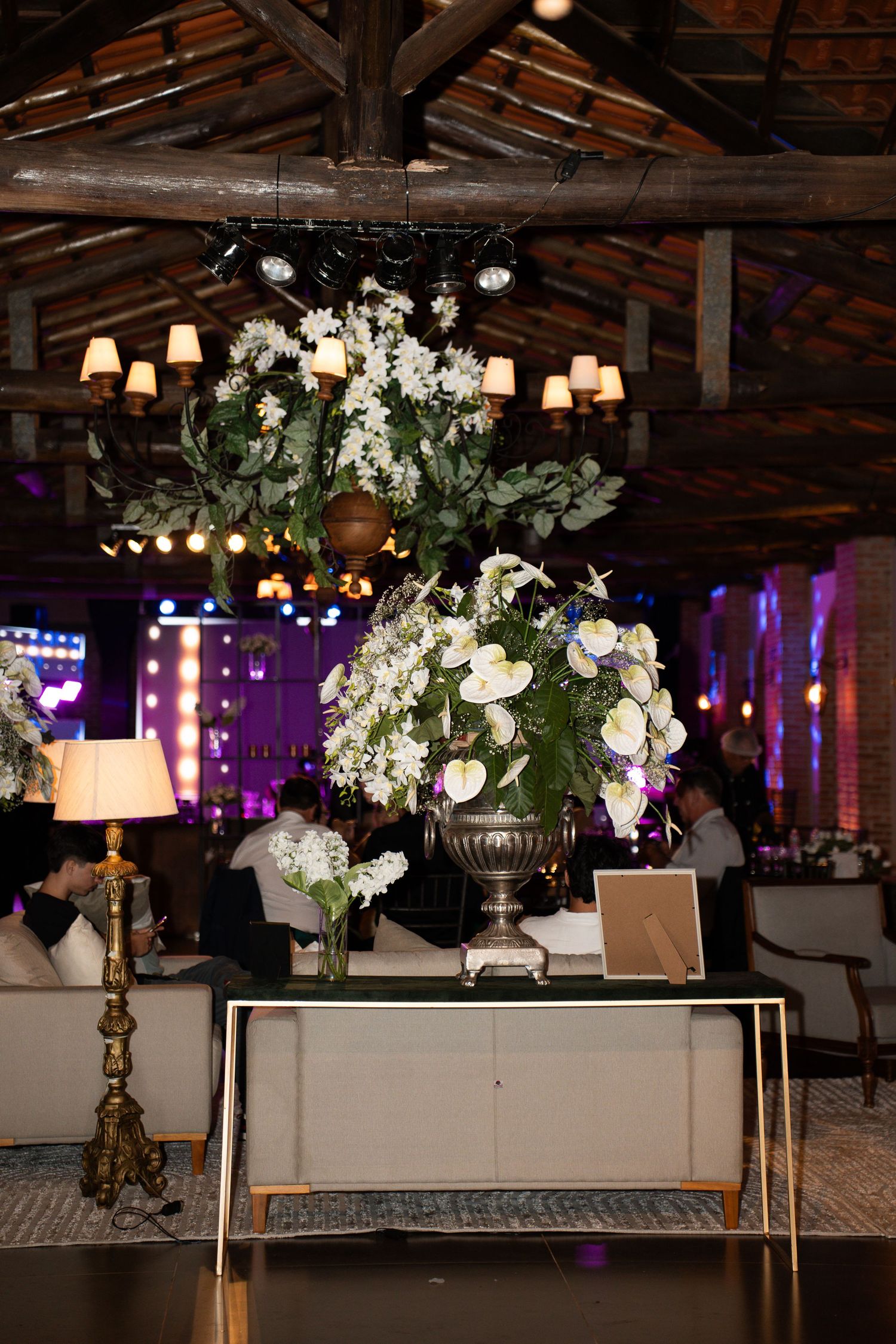 Elegant rustic wedding venue with white floral arrangements in silver urns, warm lighting, and exposed wooden beam ceiling.