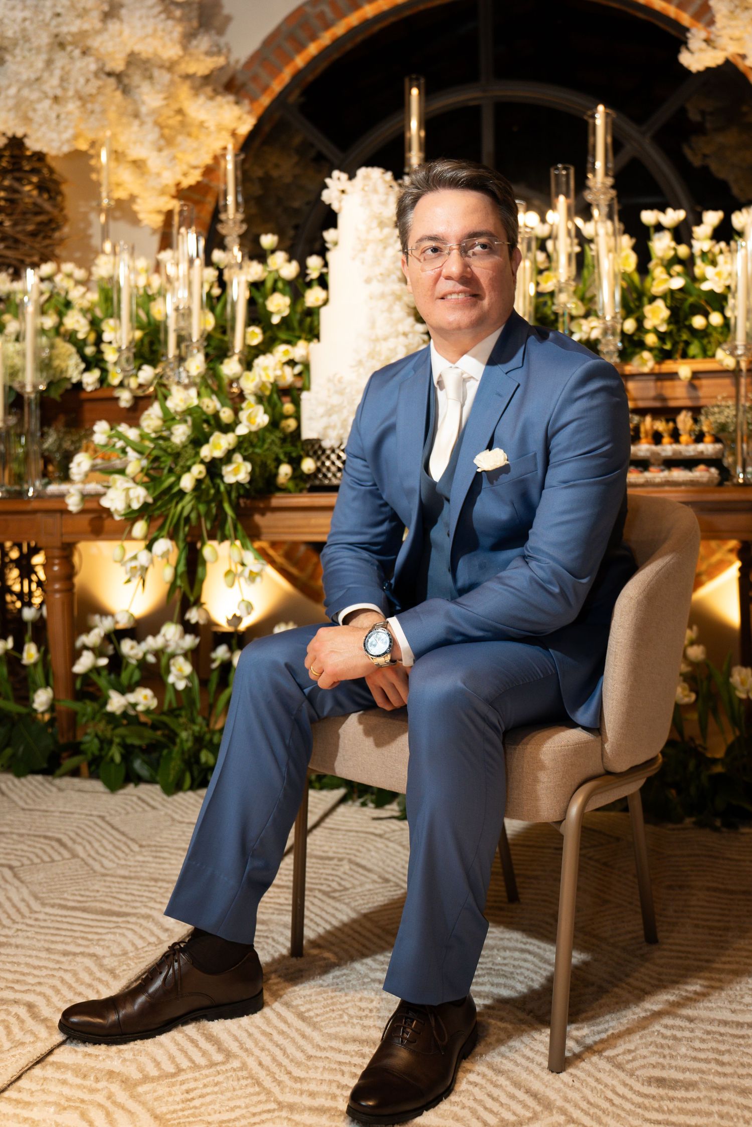 Man in blue suit sitting elegantly at a floral wedding reception backdrop with white flowers and candles.