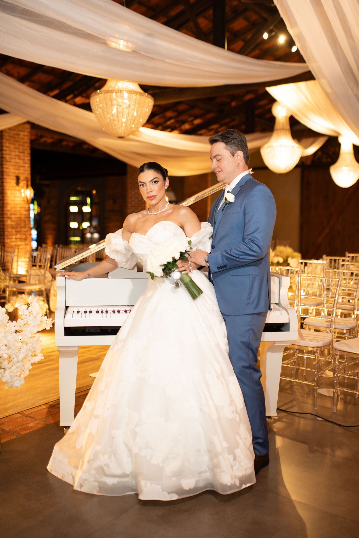 Bride in white ball gown and groom in blue suit pose by a white piano in an elegantly decorated wedding venue.