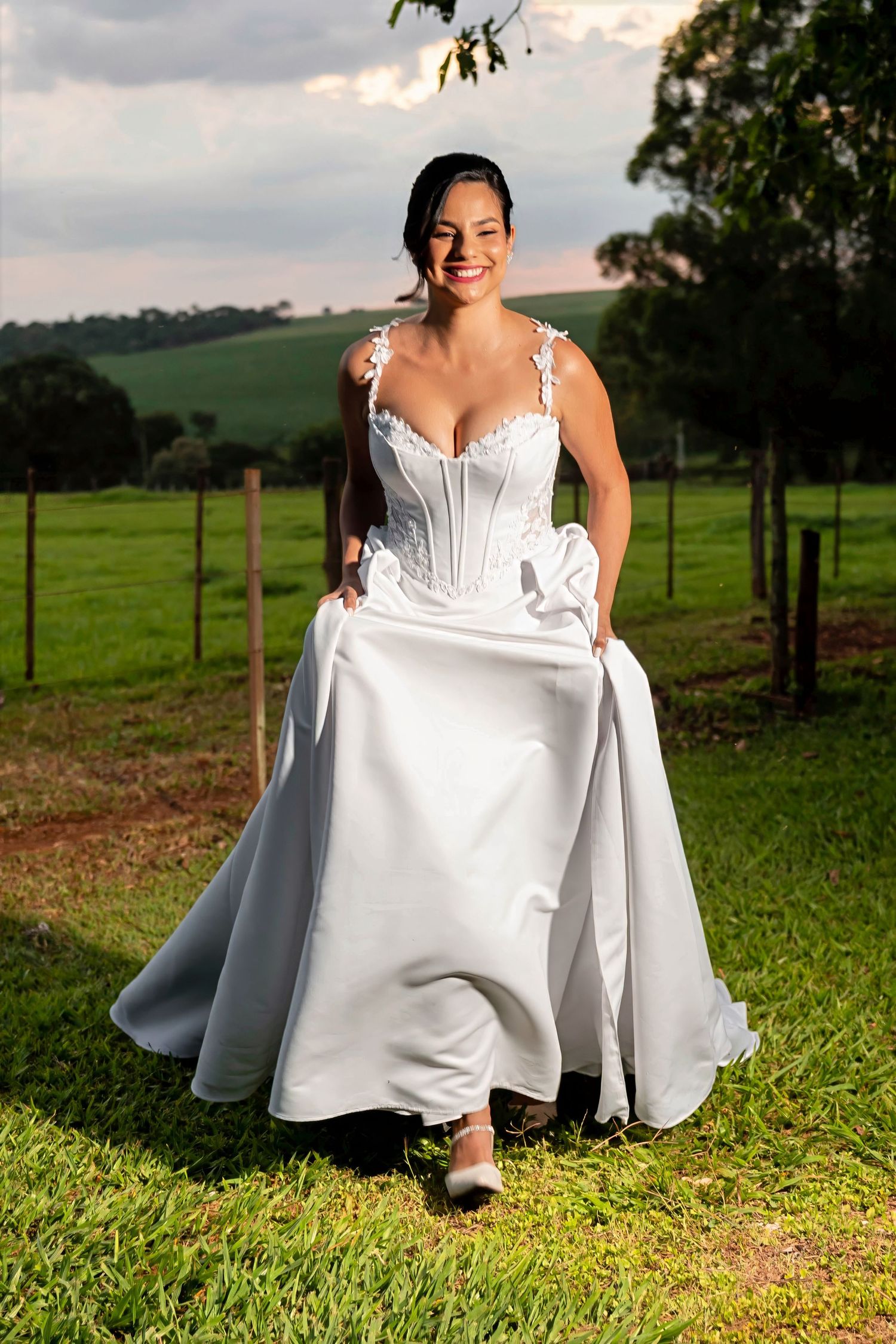 Smiling bride in a white corset wedding gown holding her skirt in a lush green countryside setting.