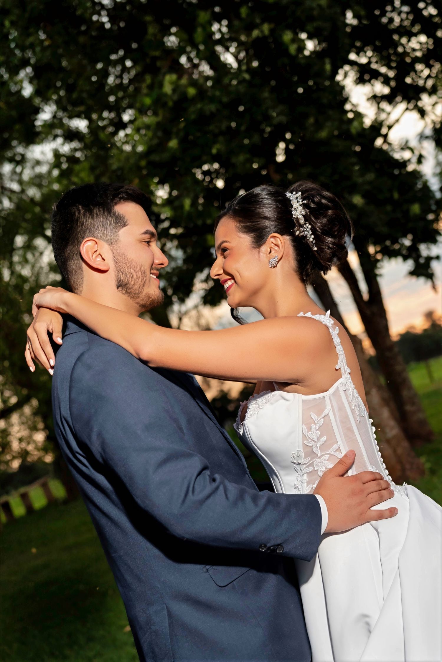 A joyful bride and groom embrace outdoors at sunset, gazing at each other lovingly in wedding attire.