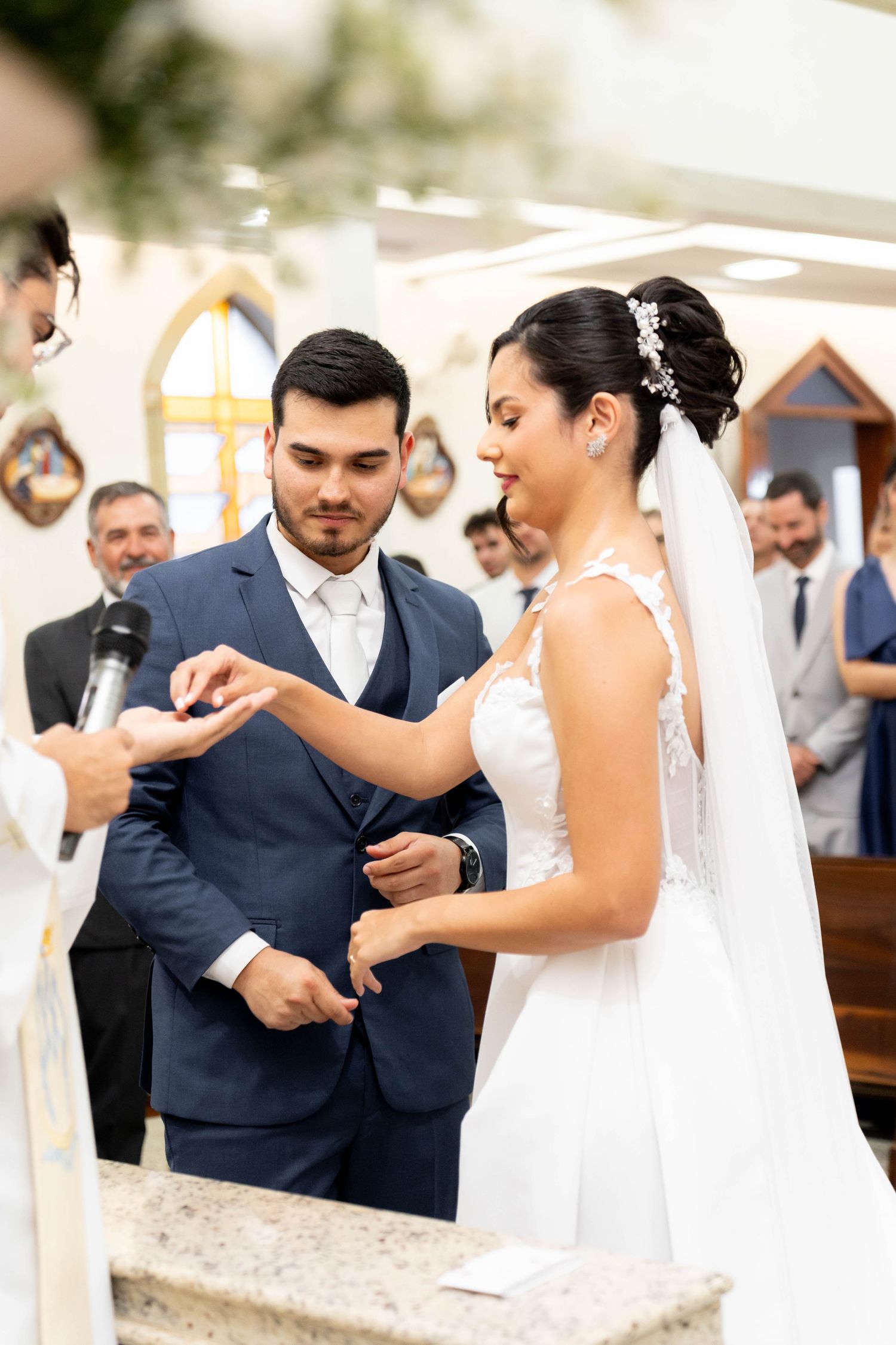 Bride placing wedding ring on groom's finger during church ceremony, both smiling joyfully.