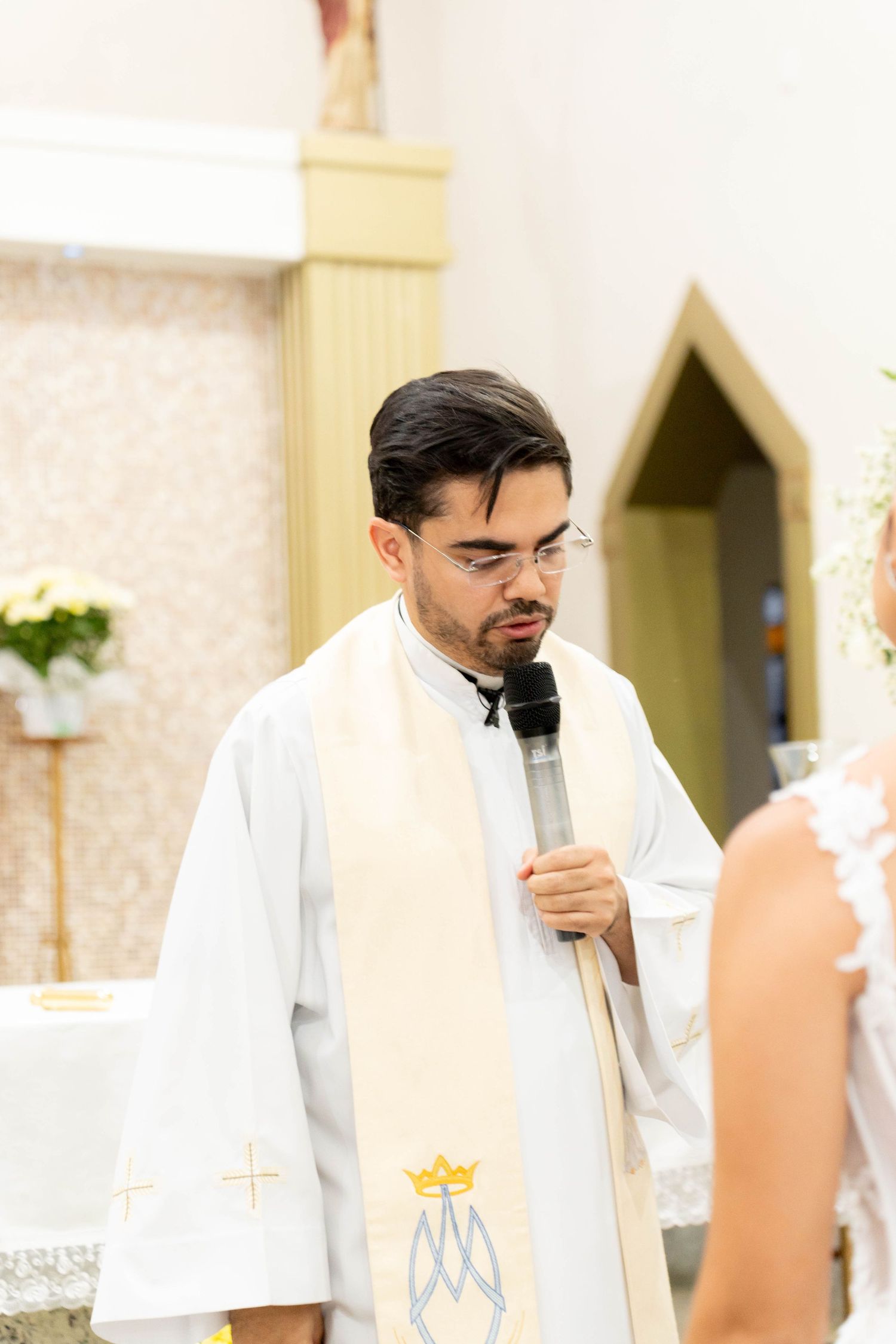 Young Catholic priest in white vestments speaks into microphone during a church ceremony at the altar.