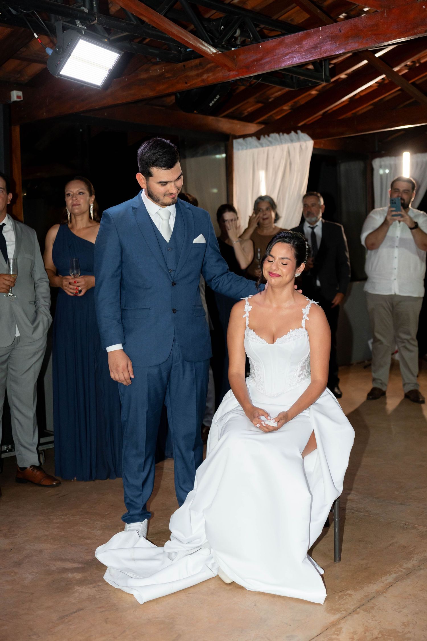 Groom in blue suit retrieves garter from bride in white wedding dress while guests watch at indoor wedding reception.