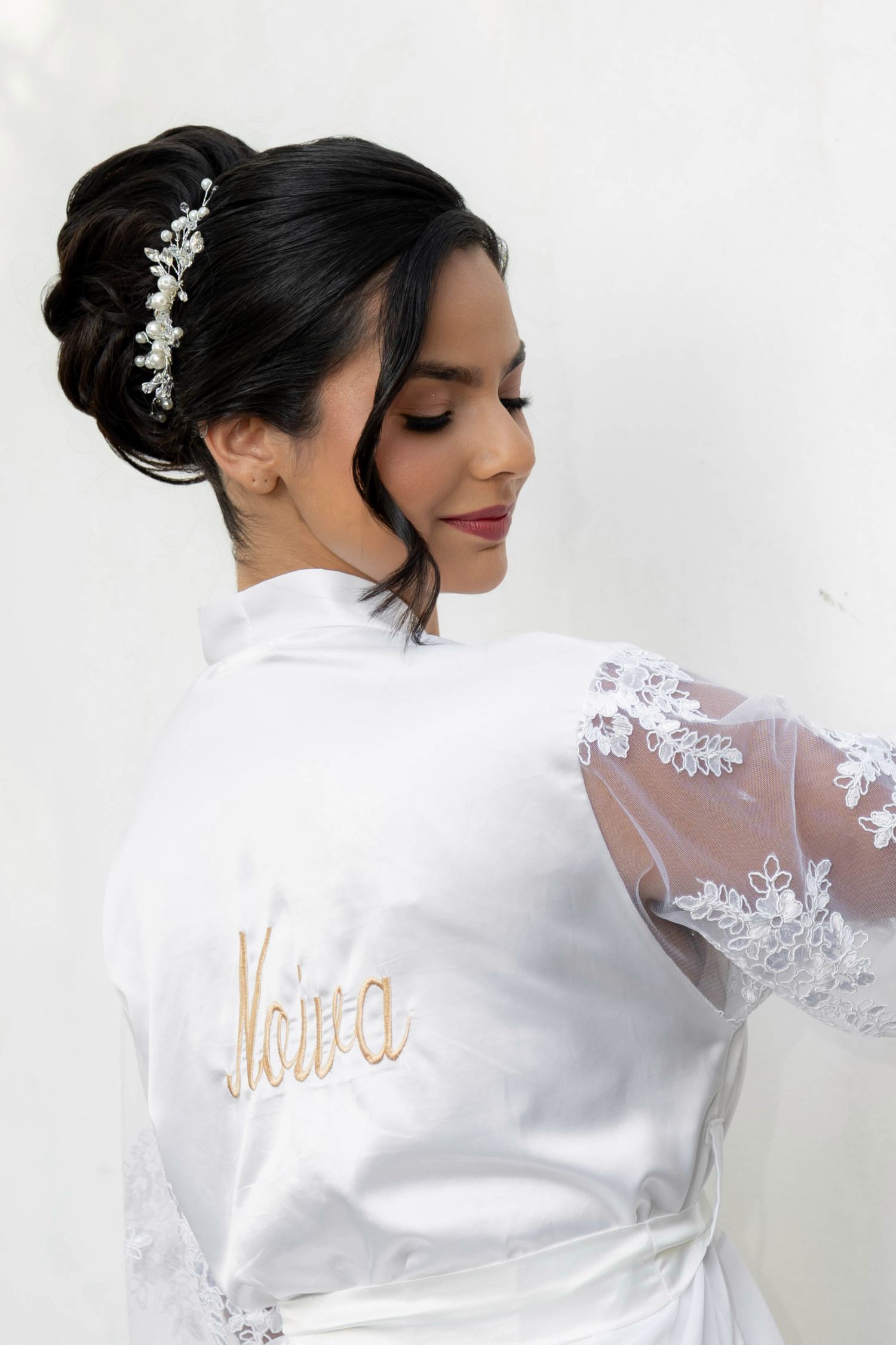 Bride in white satin robe embroidered with Noiva, wearing elegant updo with pearl hair accessory.