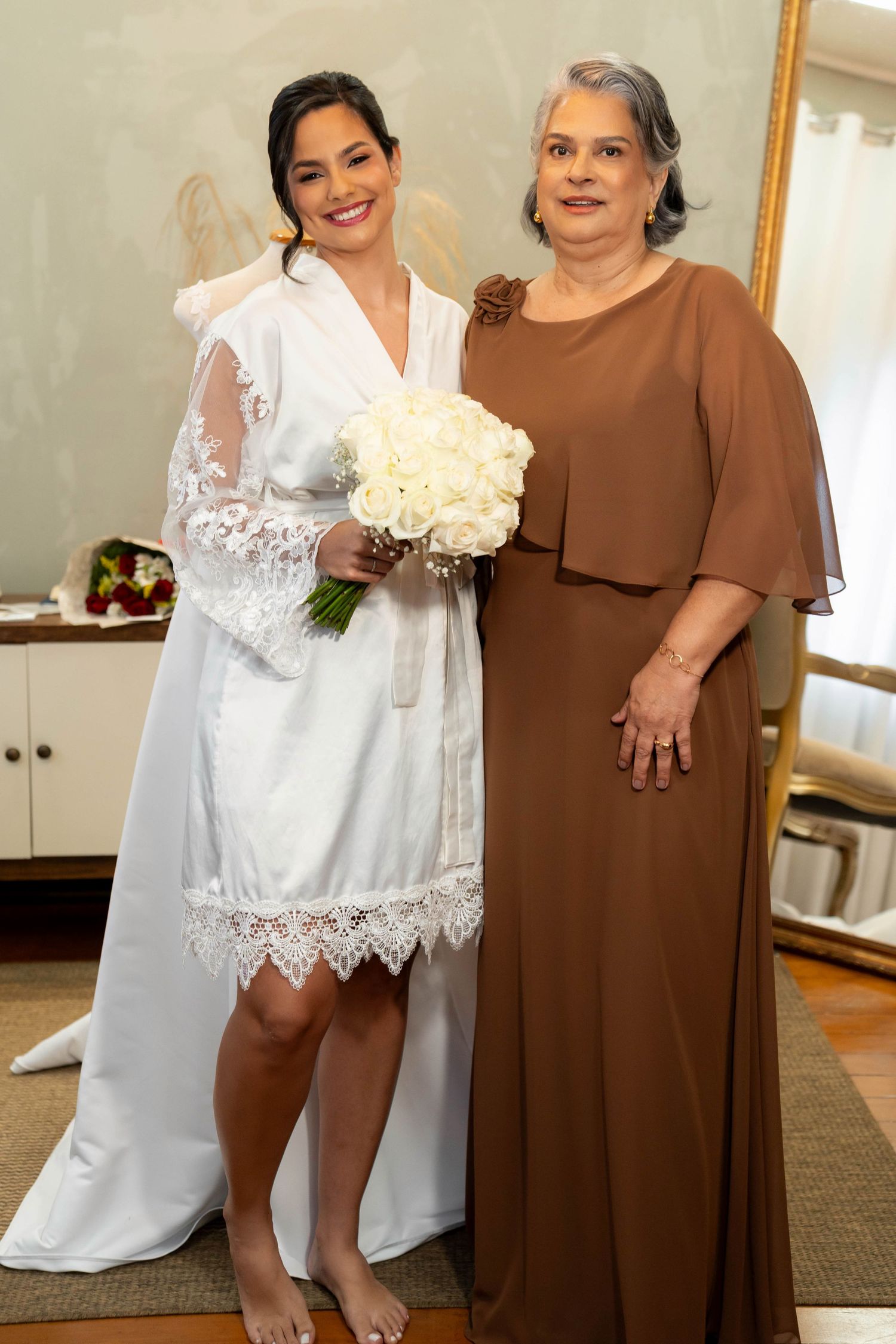 Bride in white lace robe holding bouquet of white roses stands with older woman in brown gown indoors.