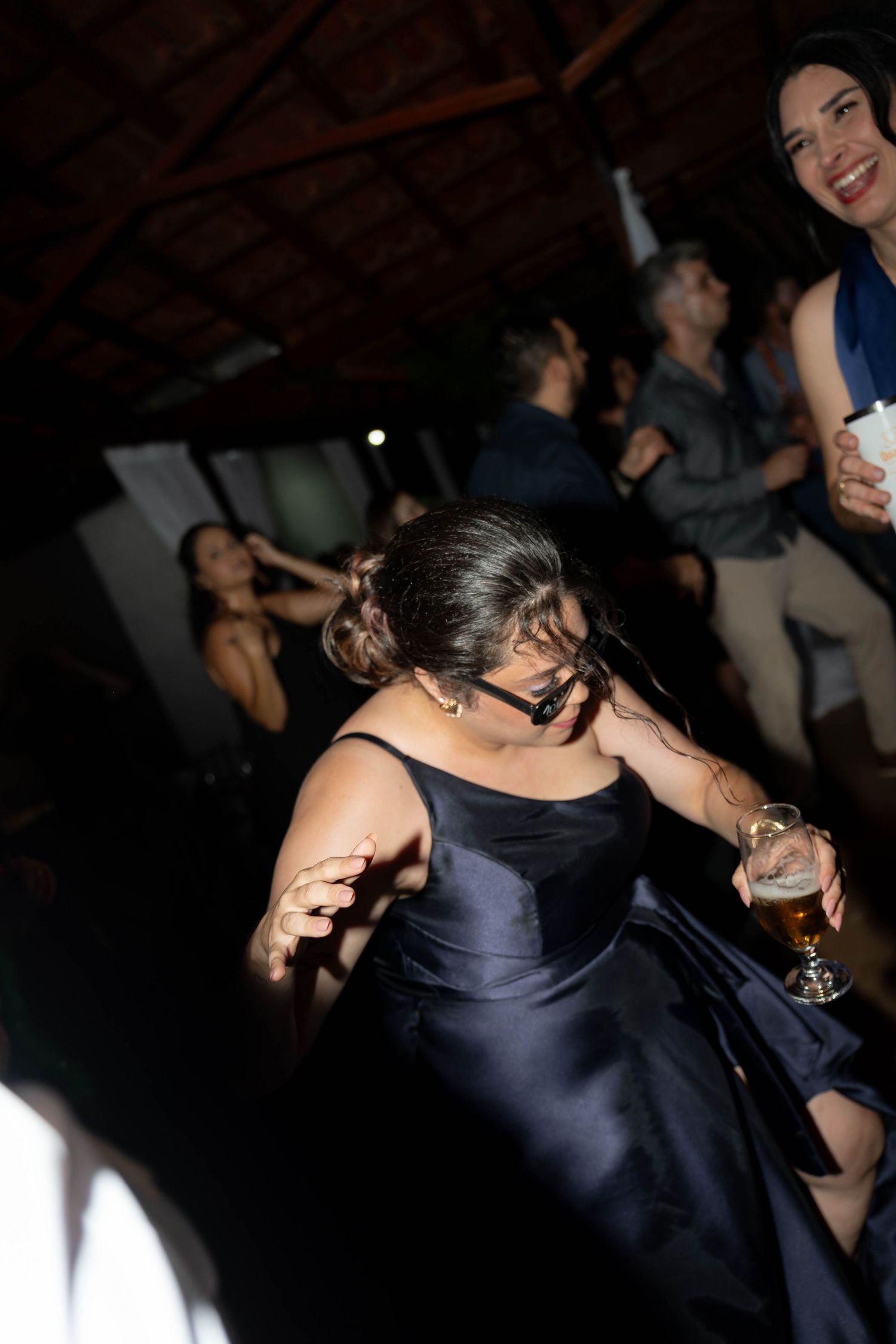 Woman in navy blue gown dancing energetically at a party, holding a drink while guests celebrate in the background.