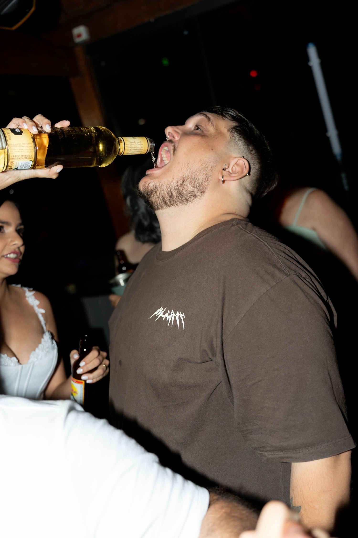 Man in brown t-shirt tilting beer bottle to drink at a nightclub party.