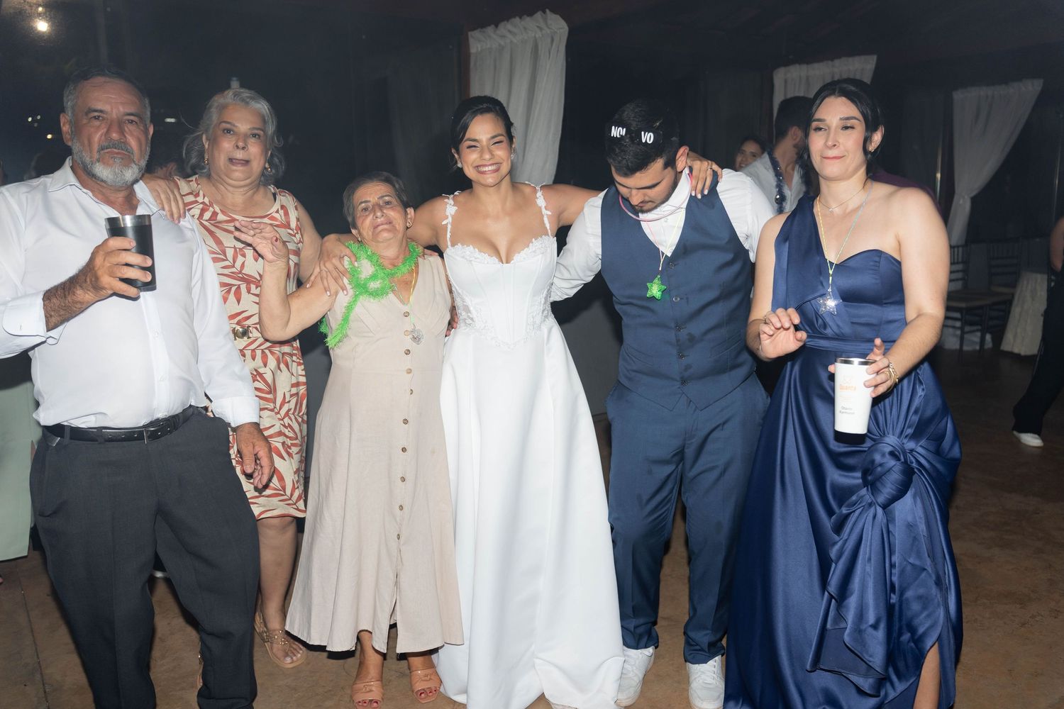 Wedding group photo with bride in white gown, groom in blue suit, and guests posing together at an outdoor reception.
