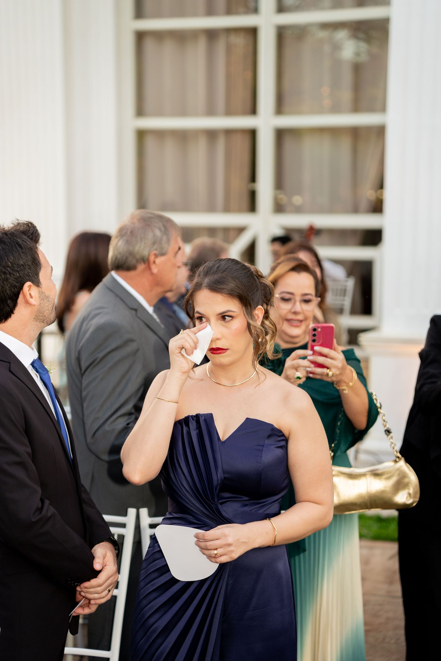 Woman in navy blue satin gown wiping tears at elegant outdoor wedding ceremony.