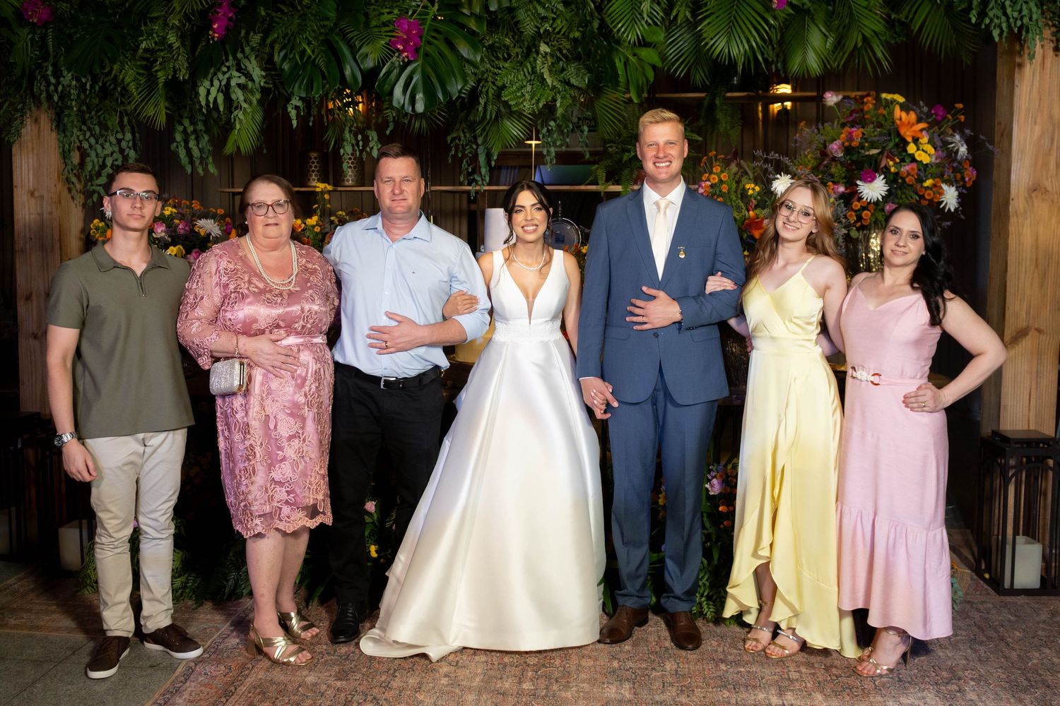 Wedding party of seven posing together indoors, bride in white gown, groom in blue suit, surrounded by family in formal attire.