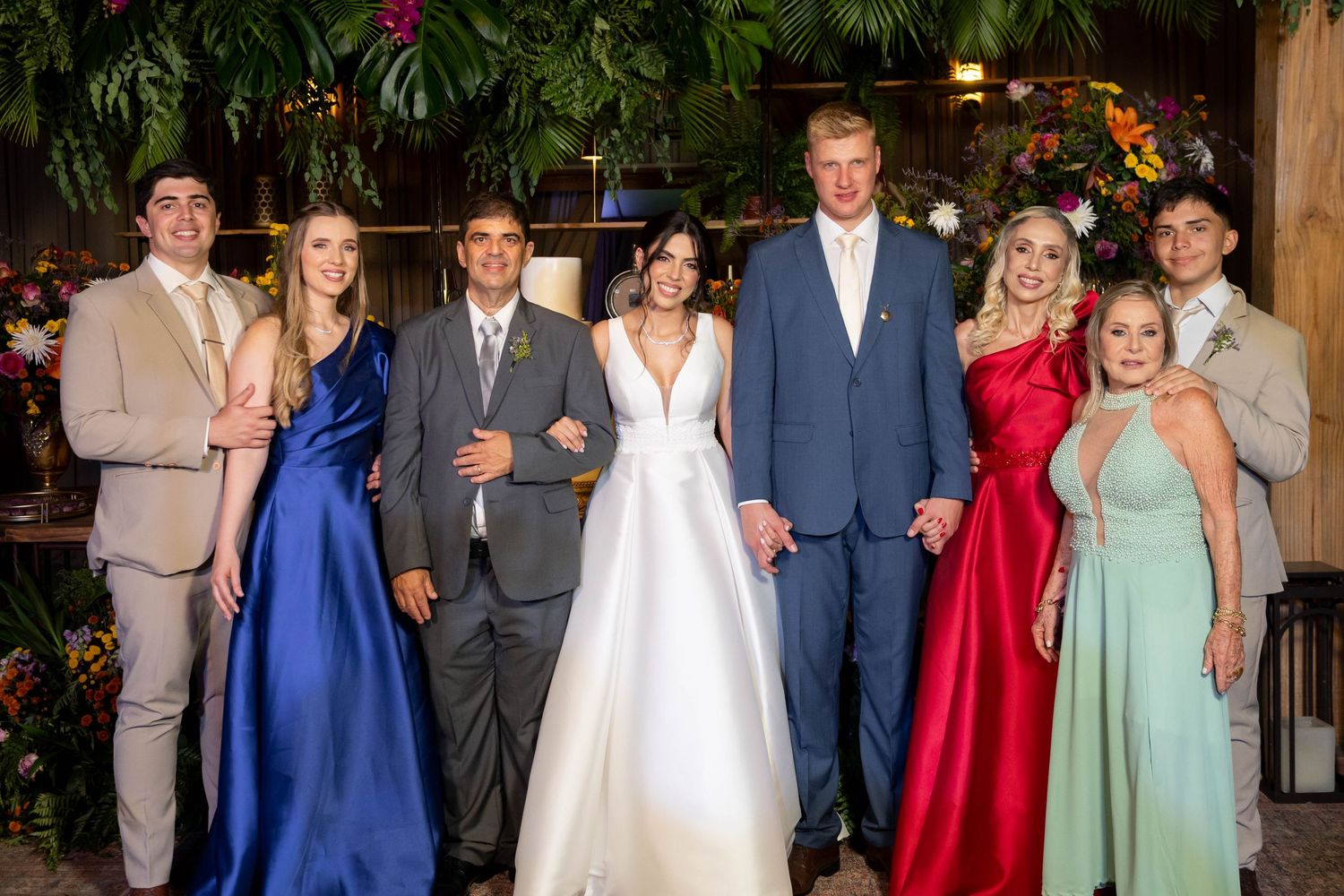 Wedding party posing together in formal attire, bride in white gown flanked by guests in blue, red, and sage green dresses.