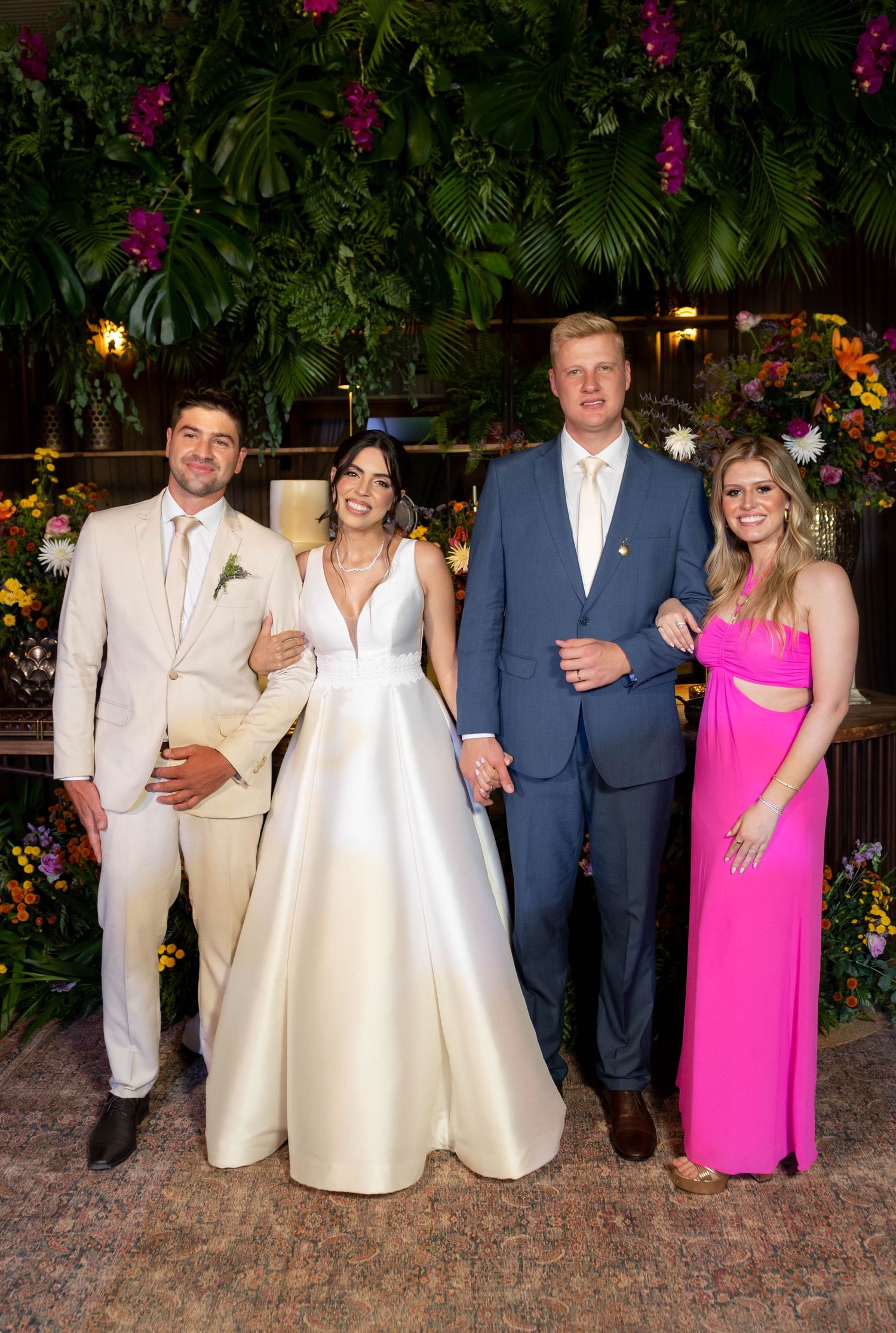 Wedding party of four posing before lush tropical floral backdrop, bride in white gown, groom in blue suit, guests in pink dress.