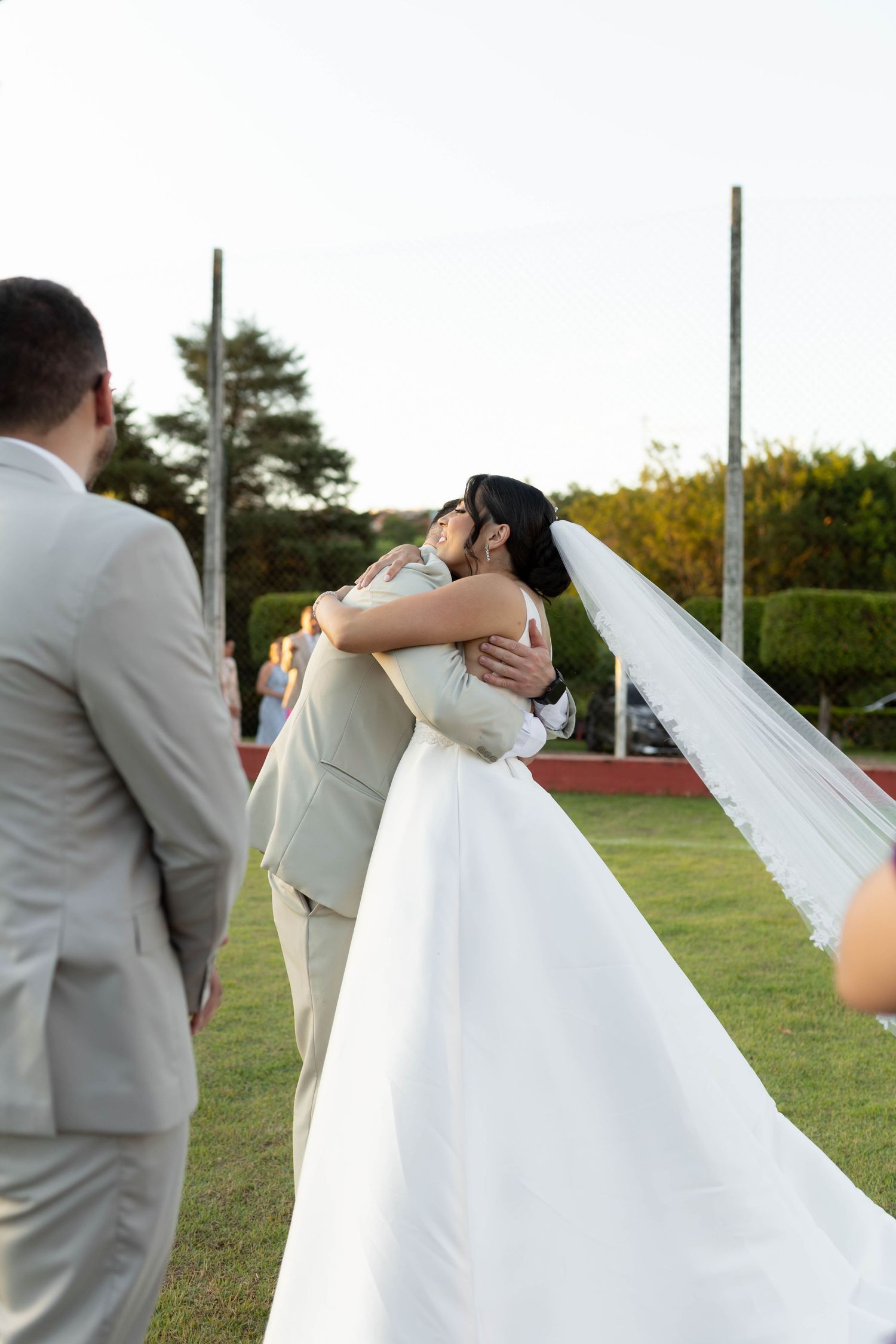 Bride and groom share a romantic embrace outdoors during their wedding ceremony, with the bride's long veil trailing behind.