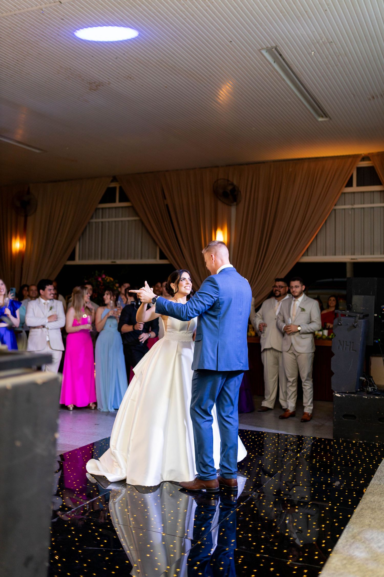 Bride and groom share their first dance on a starlit black floor while wedding guests watch in a decorated reception hall.