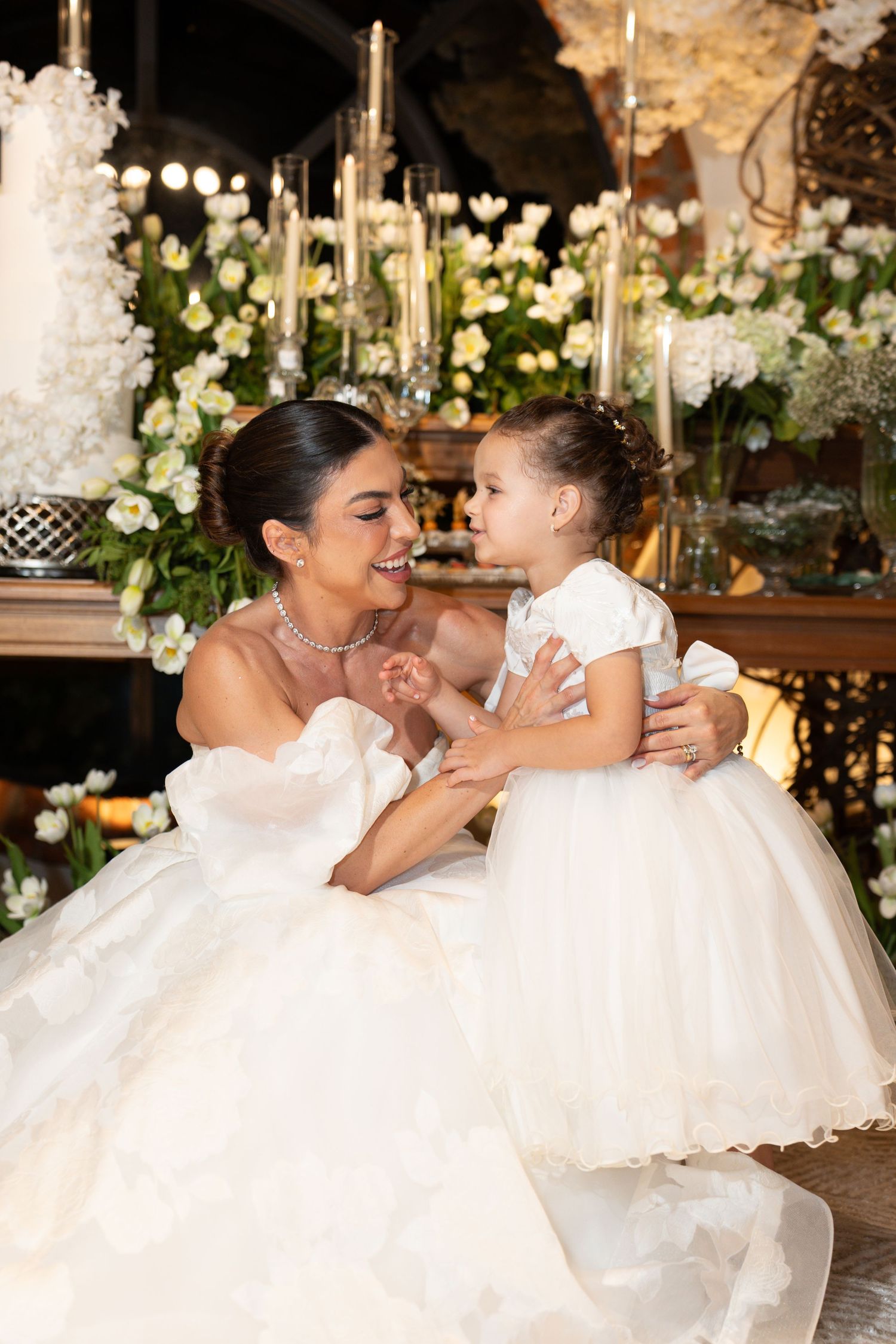 Bride in white gown smiling at flower girl in matching white dress at elegant wedding reception.