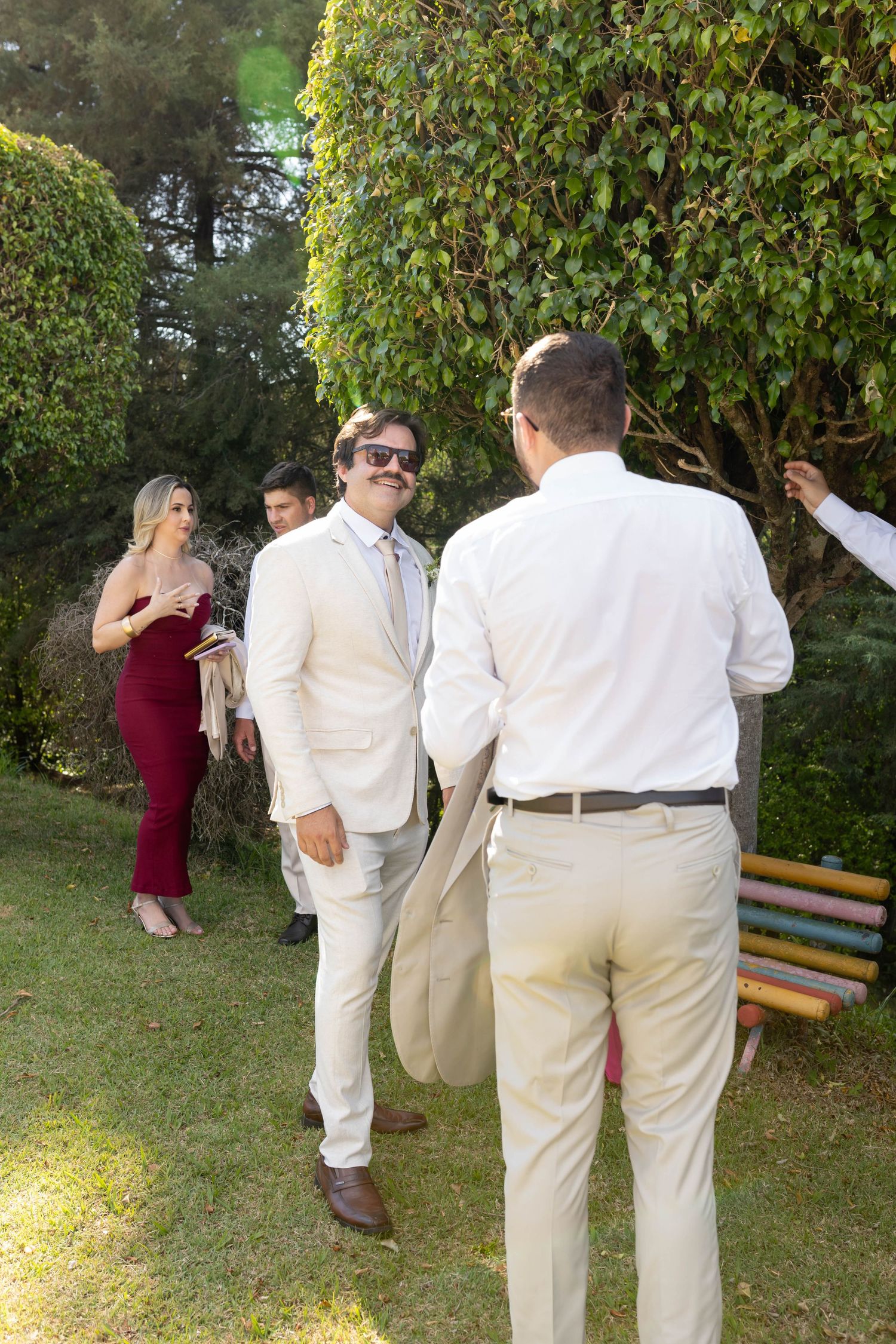 Men in beige suits chat outdoors at a garden wedding reception, with a woman in a burgundy dress visible in the background.