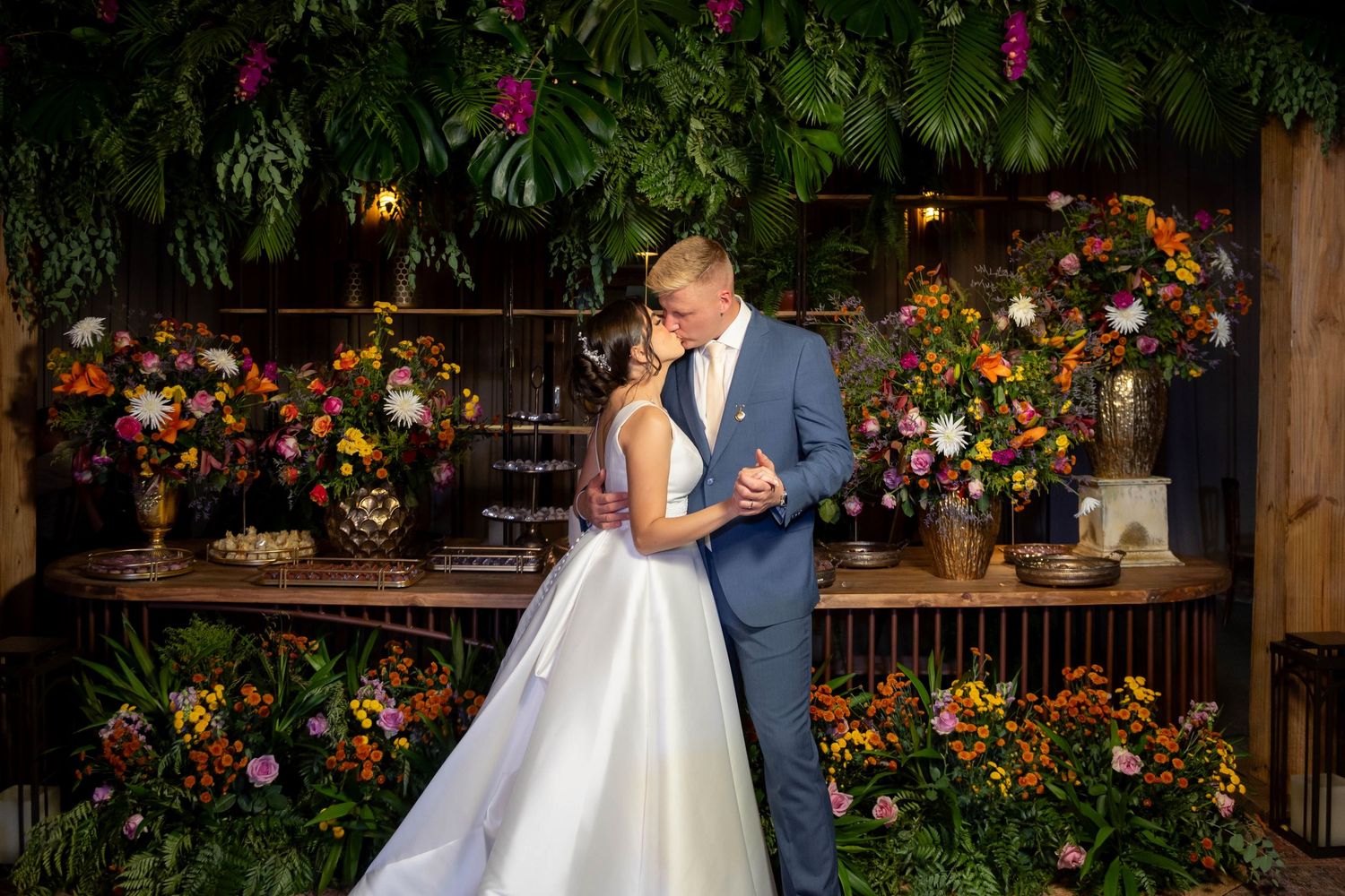 Bride and groom share romantic embrace surrounded by lush tropical greenery and vibrant floral arrangements at wedding reception.