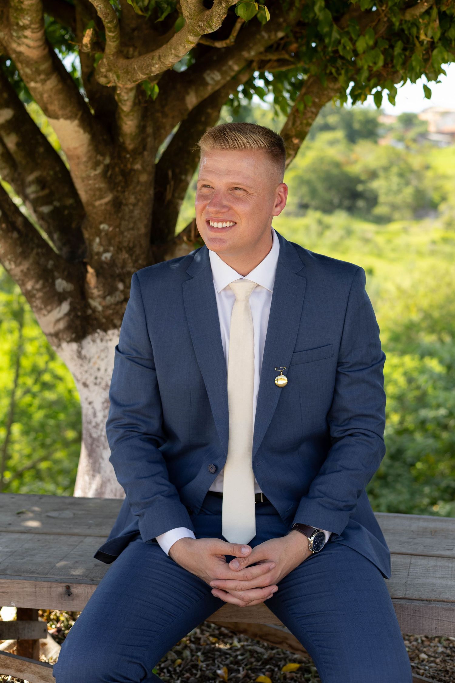 Young man in navy blue suit and white tie smiling while seated on bench outdoors under green tree.
