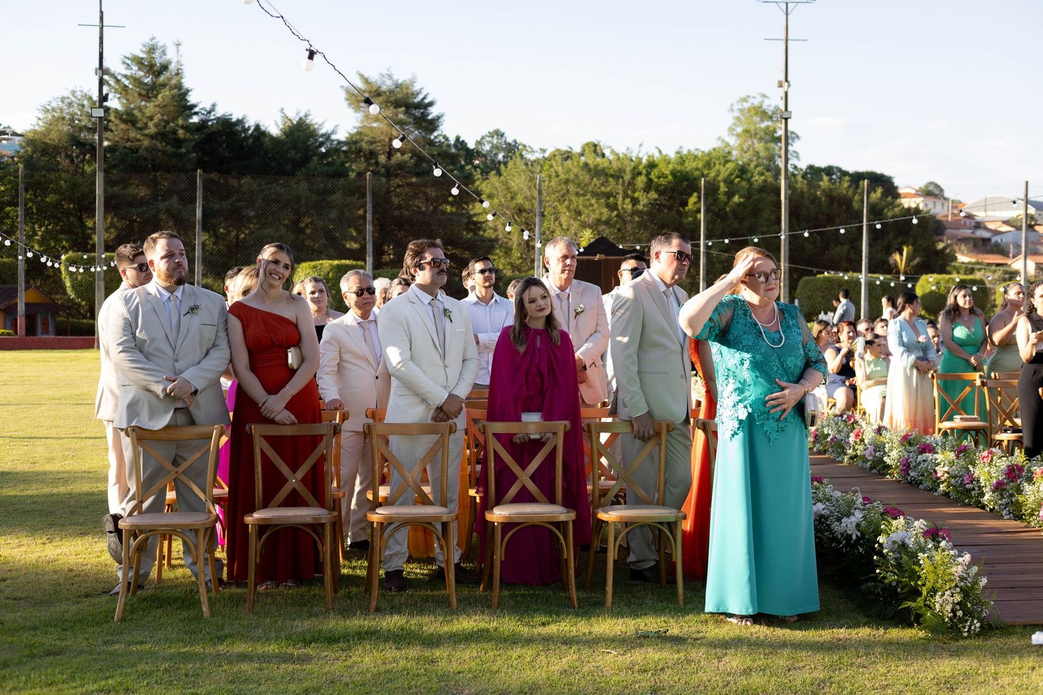 Guests in formal attire stand and sit on wooden chairs at an outdoor wedding ceremony on a green lawn.